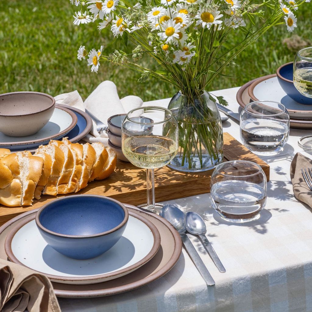 An outdoor table set for a meal - complete with flowers in a glass vase, table setting with neutral, white, and blue plates and bowls, wine glasses, sliced bread on a cutting board, and steel flatware.