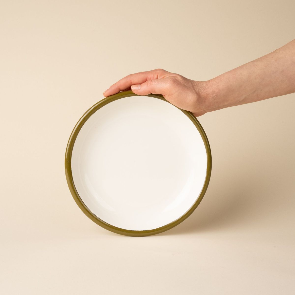 A hand props up an enamel side plate in white with an olive green rim.
