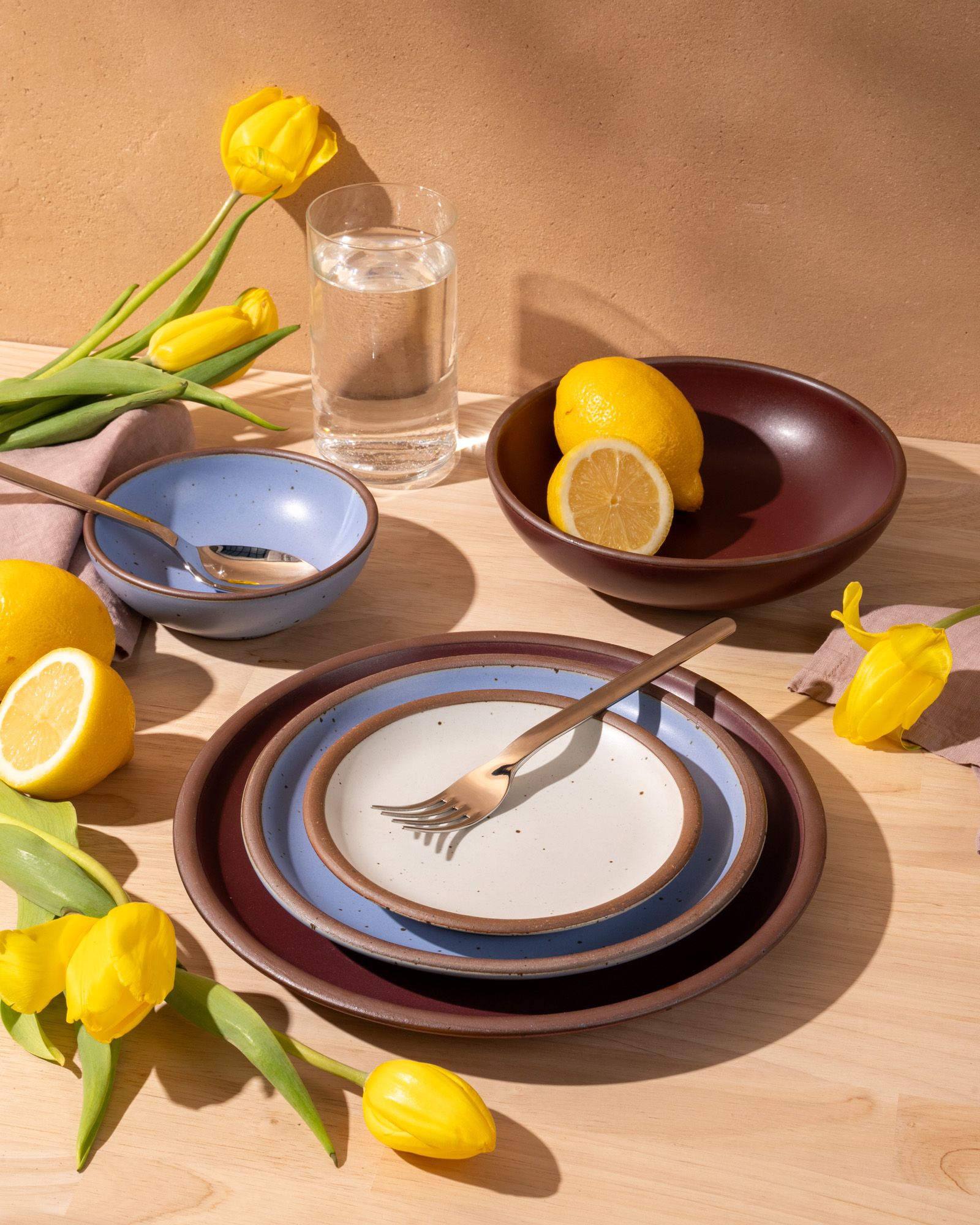 An artful dinnerware set featuring 3 different sized plates and 2 small and large shallow bowls in plum, periwinkle and cool white colors on a table. There is also silverware on the plates, a tall glass of water, and adorned yellow tulips and lemons.