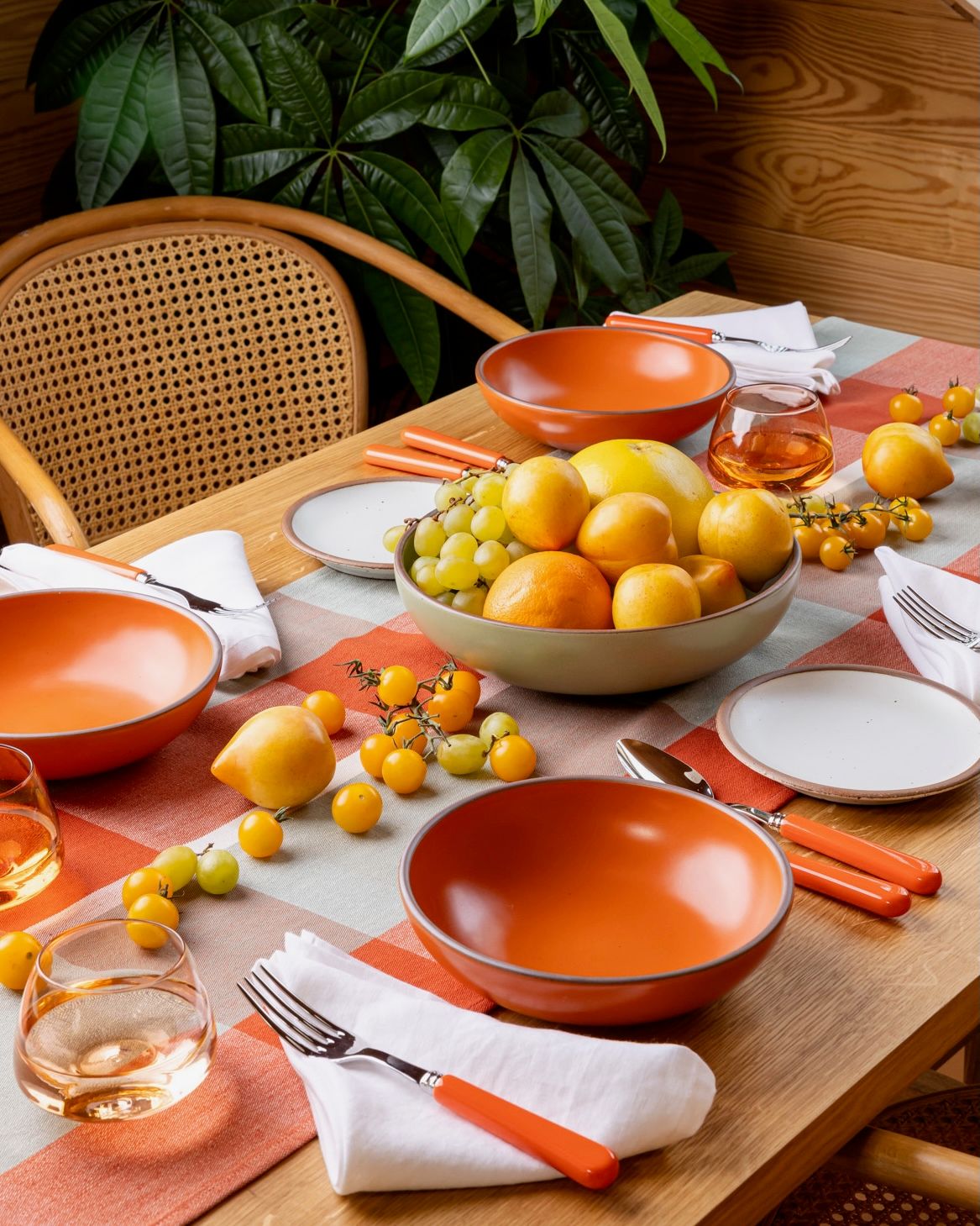 A table filled with several place settings, featuring a shallow dinner bowl in a bold orange, a folded white napkin, orange flatware, whiskey snifters, and a centerpiece filled with fruits and vegetables, all in shades of yellow.