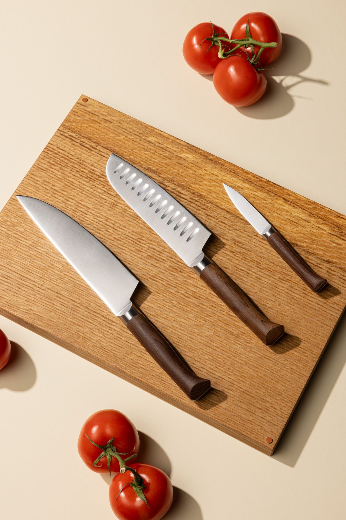 Three different kitchen knives lined up on a wooden cutting board with tomatoes surrounding. The knives are a chef's knife, a santoku knife, and a paring knife.