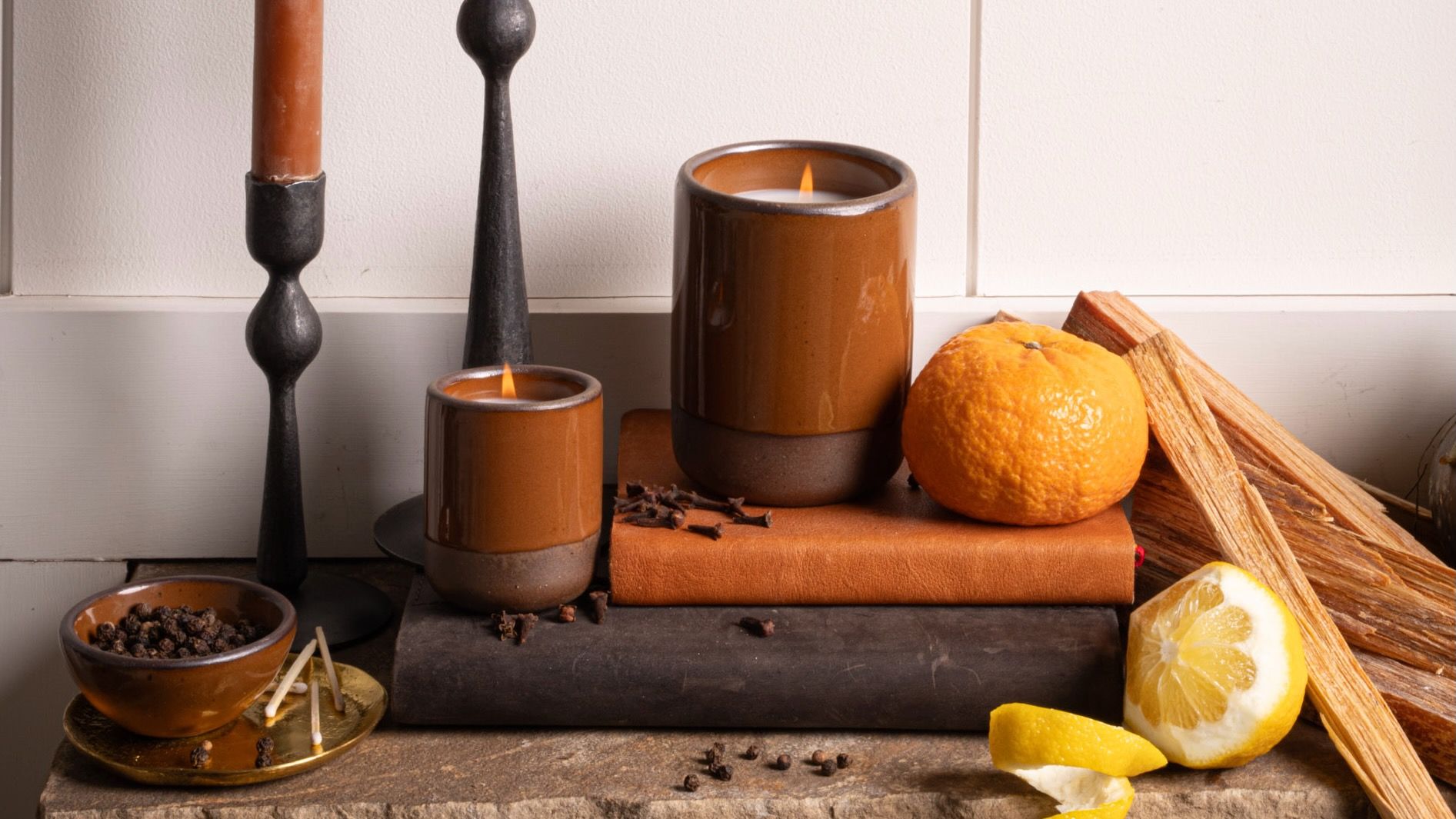 Two brown glazed candles burn beside citrus, spices, and palo santo on a stone mantle, with tall metal candlesticks in the background.