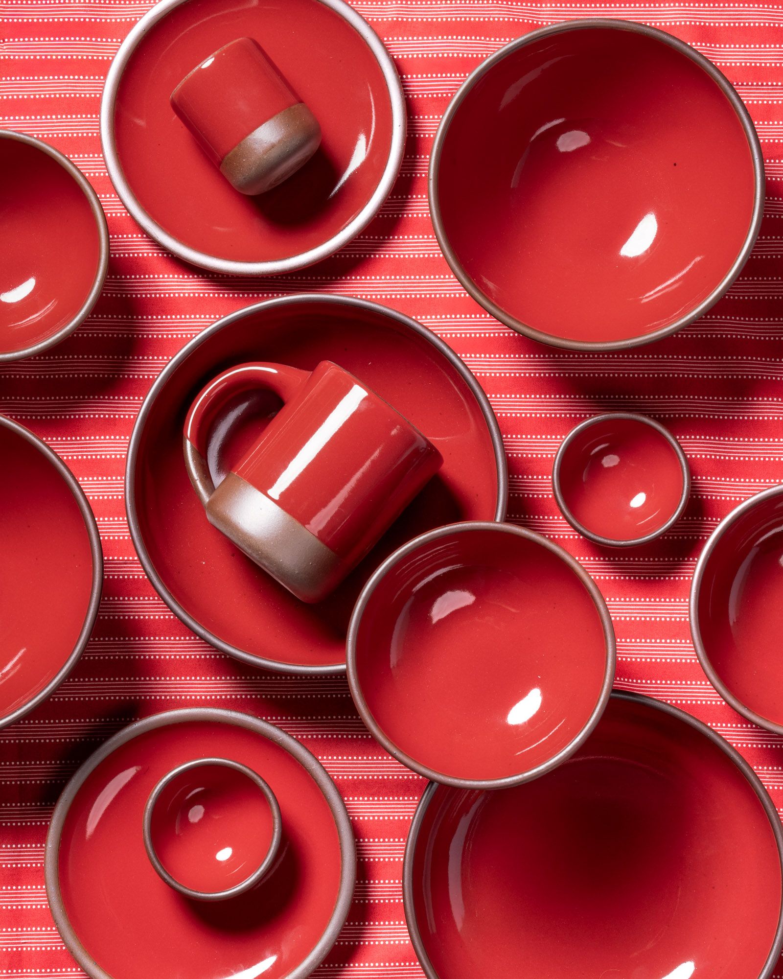 An overhead view of glossy red ceramic bowls, mugs, and plates against a red striped tablecloth.