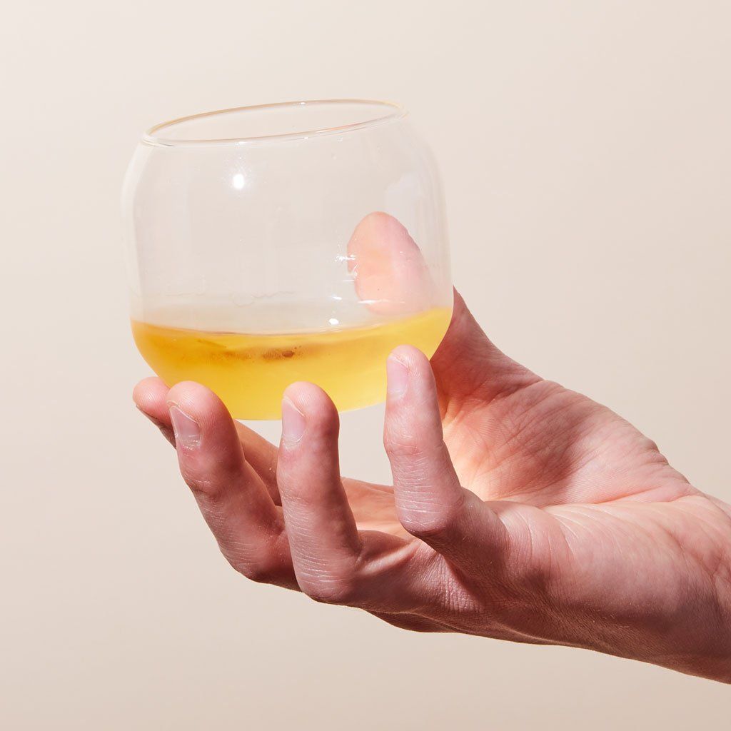 Yellow liquid in a rounded, stemless wine glass held by the fingers and thumb of a hand