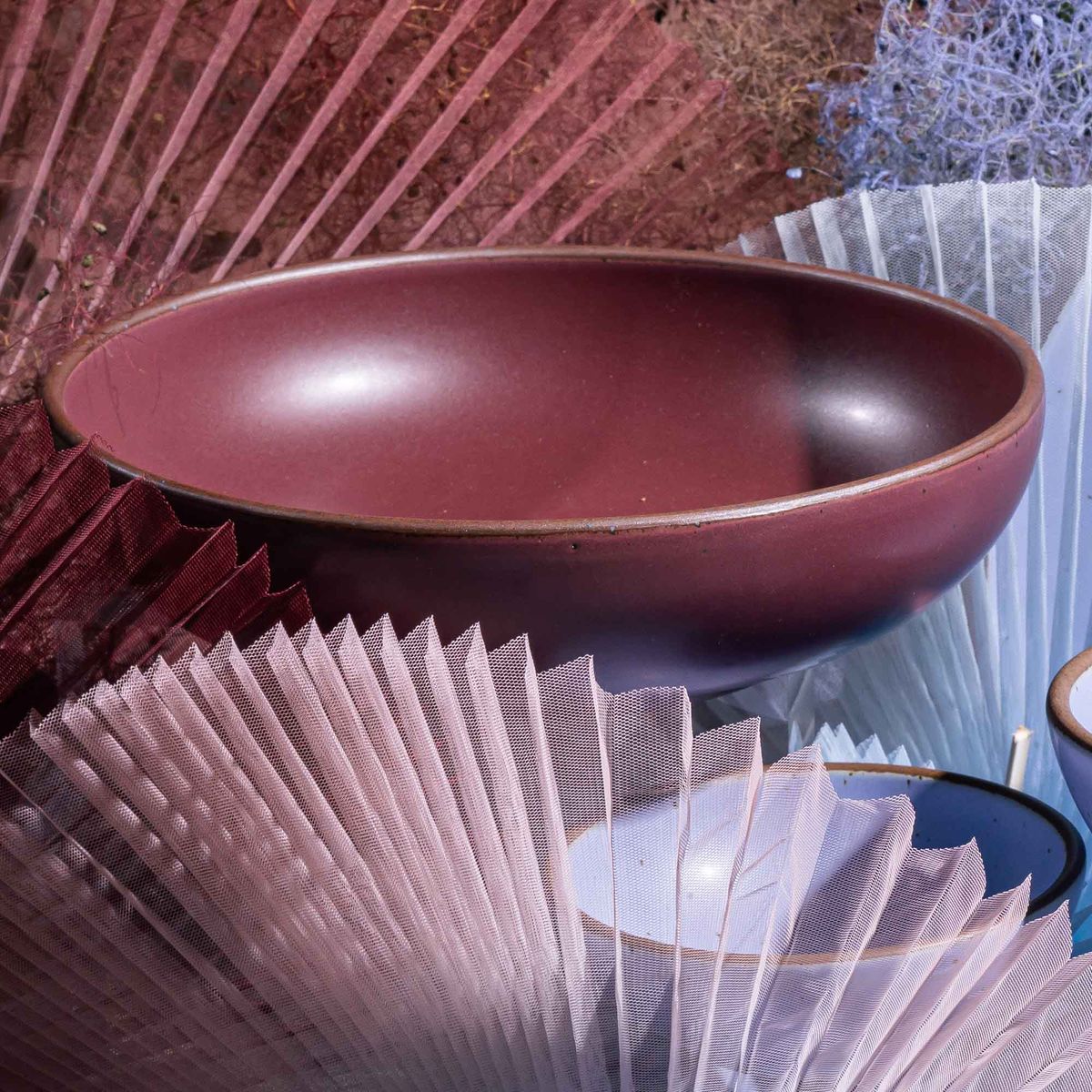 A large shallow serving ceramic bowl in a plum color surrounded by a pleated fabric sculpture