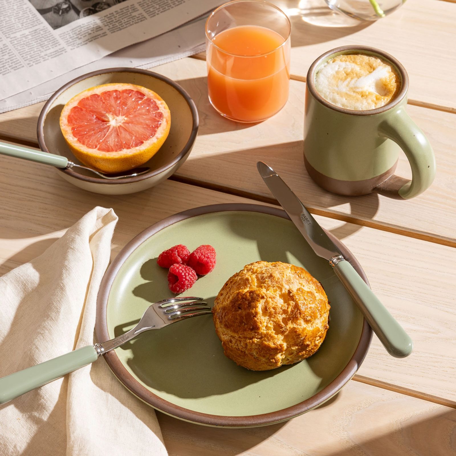 On a breakfast table, there are a ceramic plate, bowl, and mug - all in a sage green. The bowl has grapefruit, the plate has a pastry and berries, and the mug has coffee. Surrounding are a newspaper, sage green flatware, and a natural colored napkin.