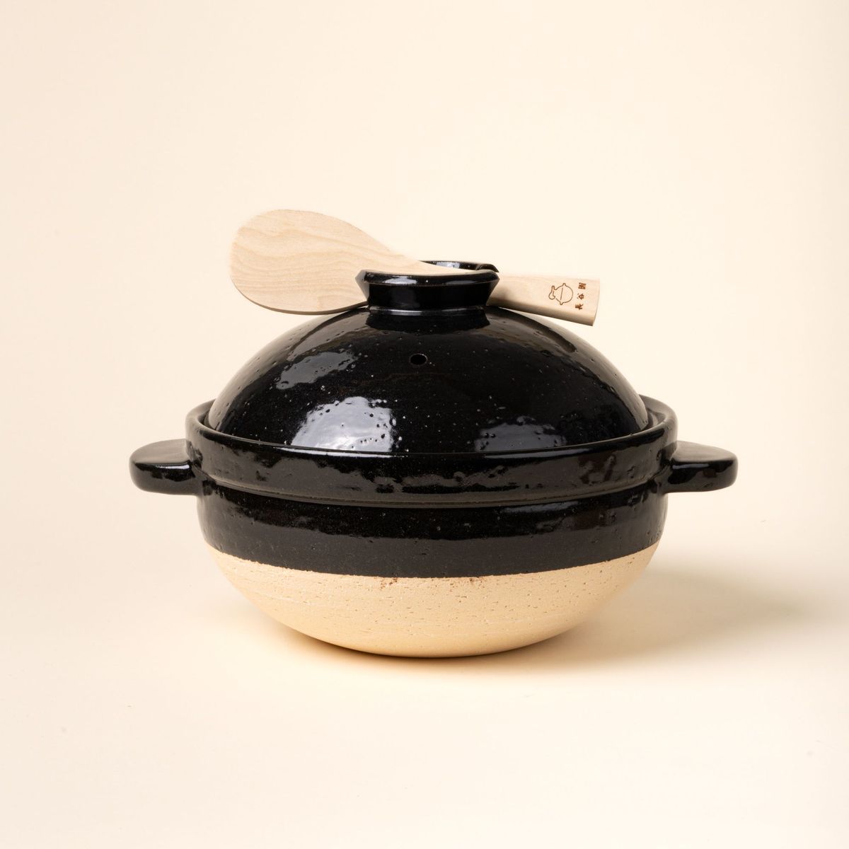 A black-and-neutral color claypot with handles and a shallow-dome shaped lid. The cooking vessel sits atop a wooden board next to a wooden spoon.