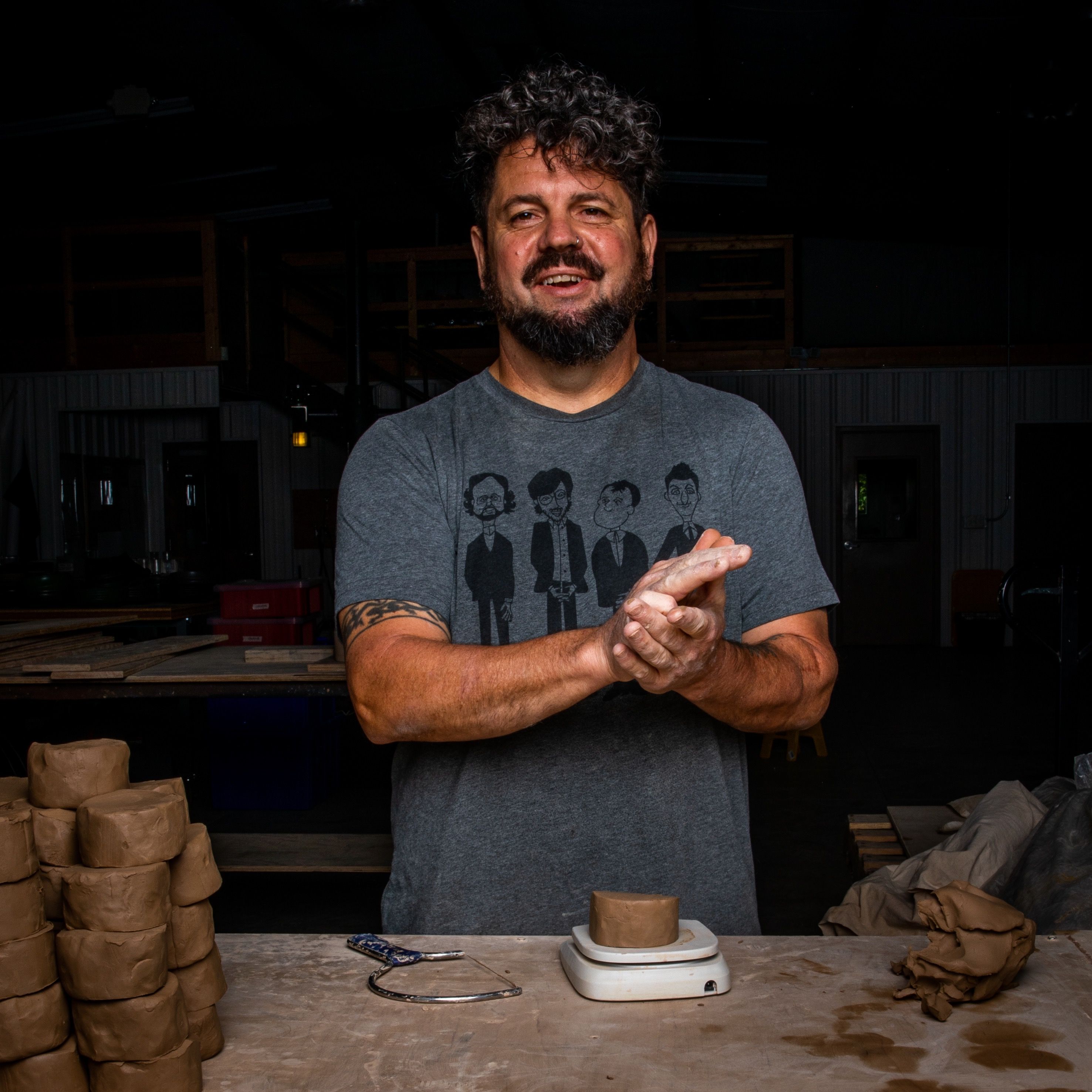 A man is staring directly at the camera and smiling. He is in a clay workshop environment and is diving pieces of clay into small batches with a clay cutter and scale.. 