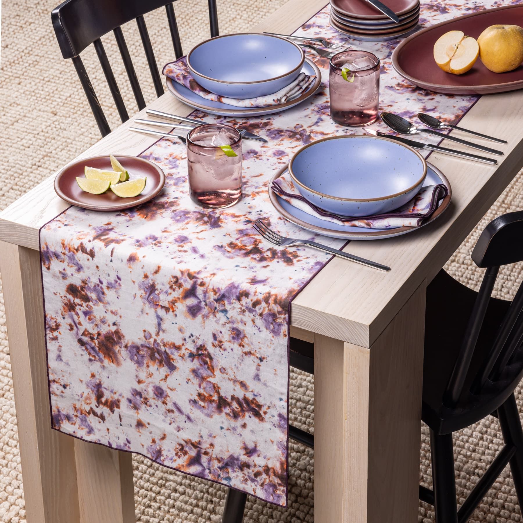 A table runner with a marble pattern featuring pink, purple, orange and white colors sits on a table with periwinkle ceramic dishes, pink glasses, and foods.