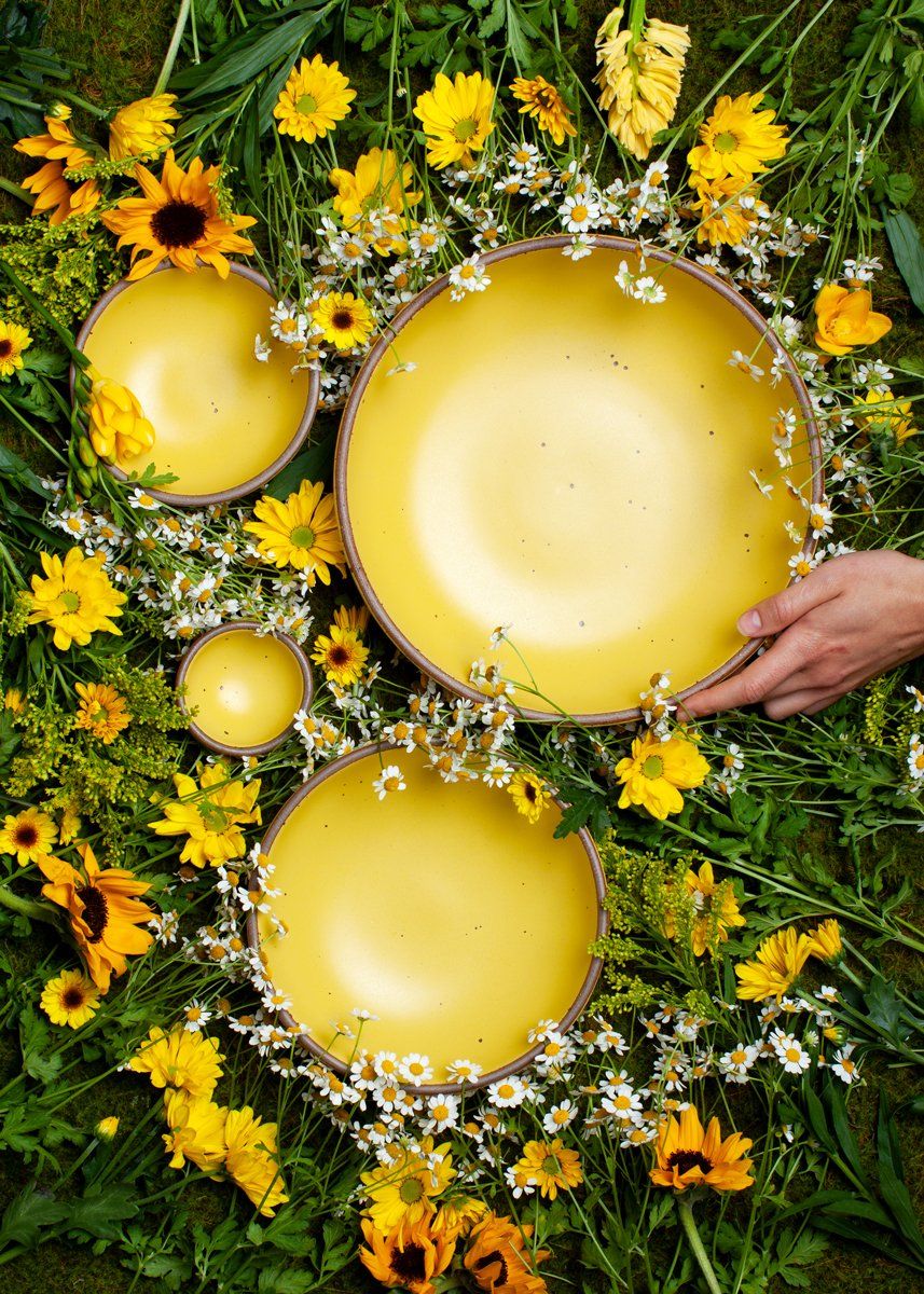 An overhead view of bright yellow ceramic dishes artfully arranged in the grass with golden and yellow flowers. A hand reaches for one of the pots.