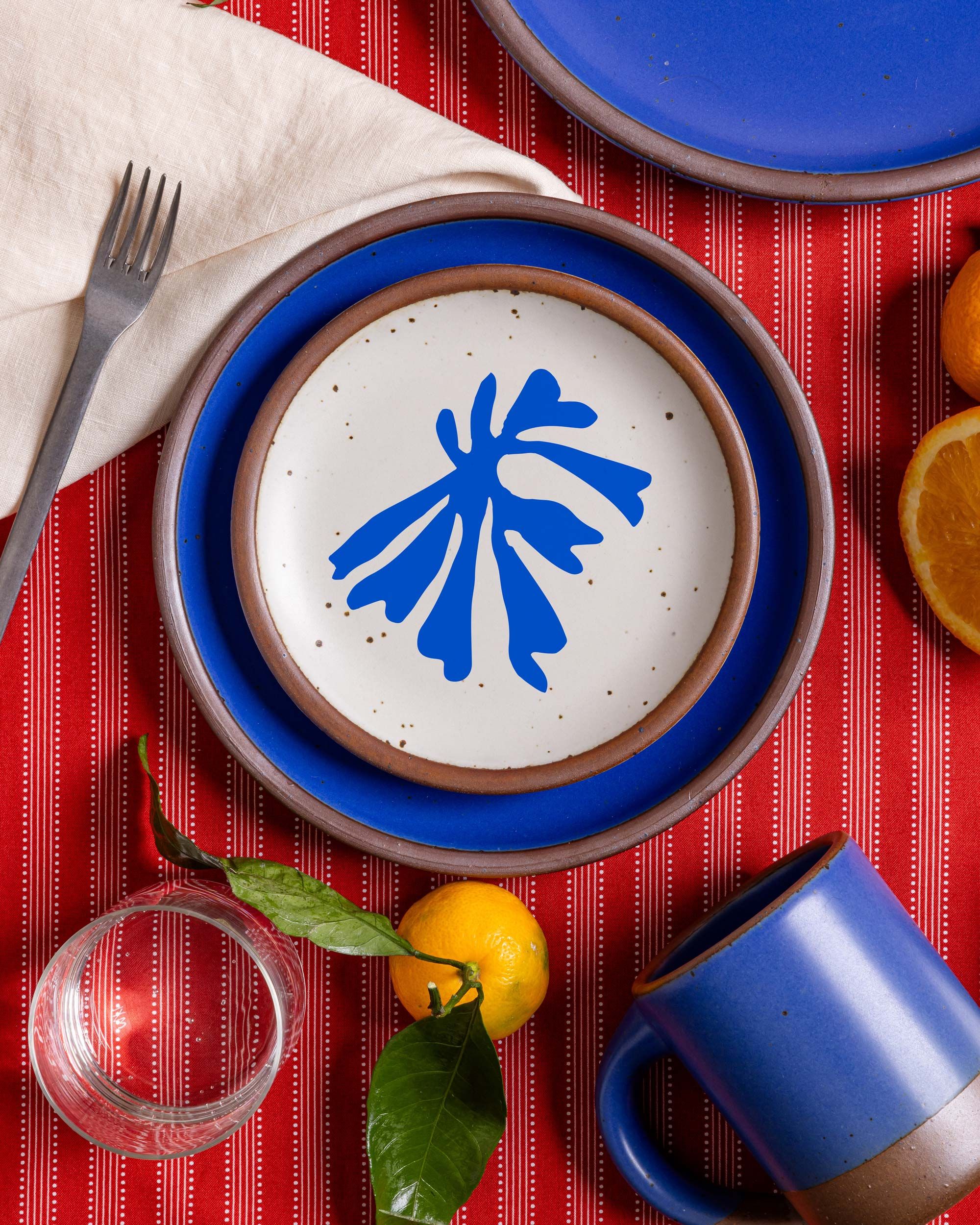 A close-up of an East Fork place setting with a Matisse Collection salad plate featuring a bold blue abstract leaf decal atop a La Sirène blue dinner plate. A deep blue mug, a folded cream napkin, a fork, glass of water, and scattered citrus fruits with leaves add texture and color to the red patterned tablecloth.