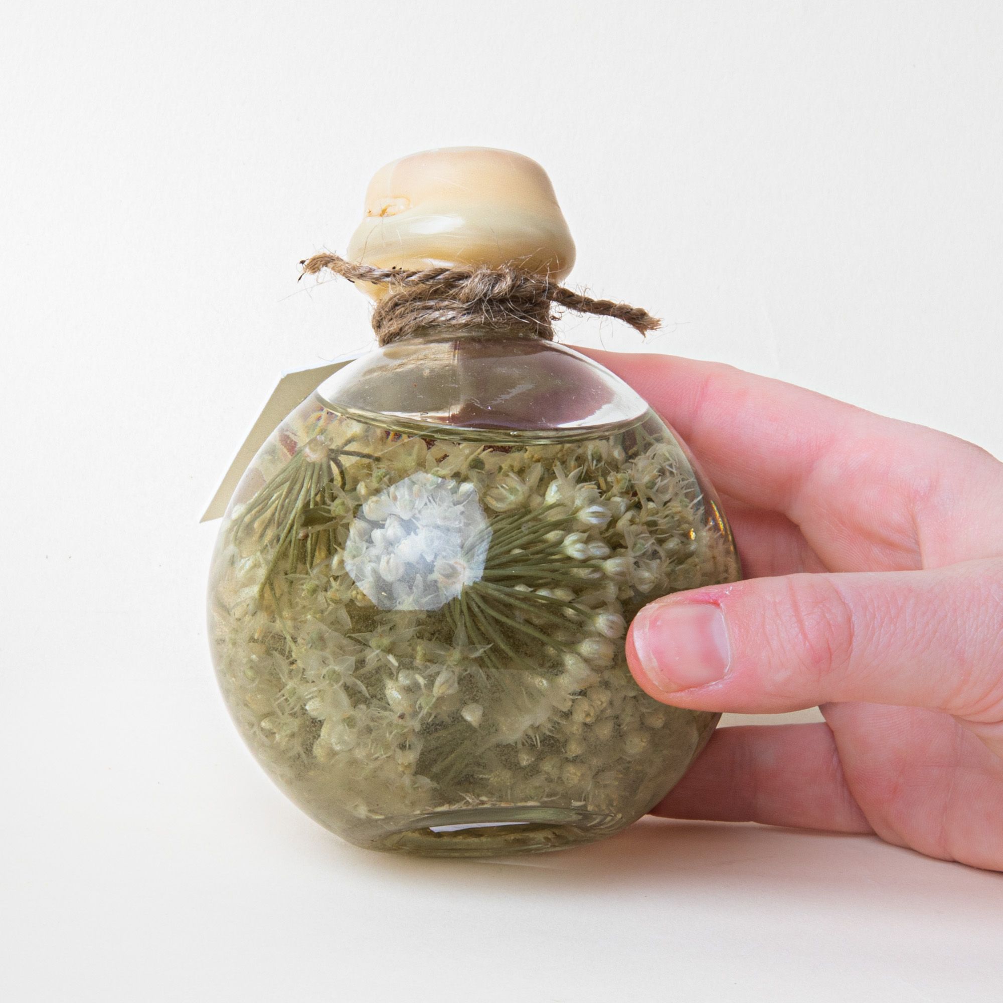 A hand holding a round jar filled with flowery liquid in a sage green color with a wooden cap