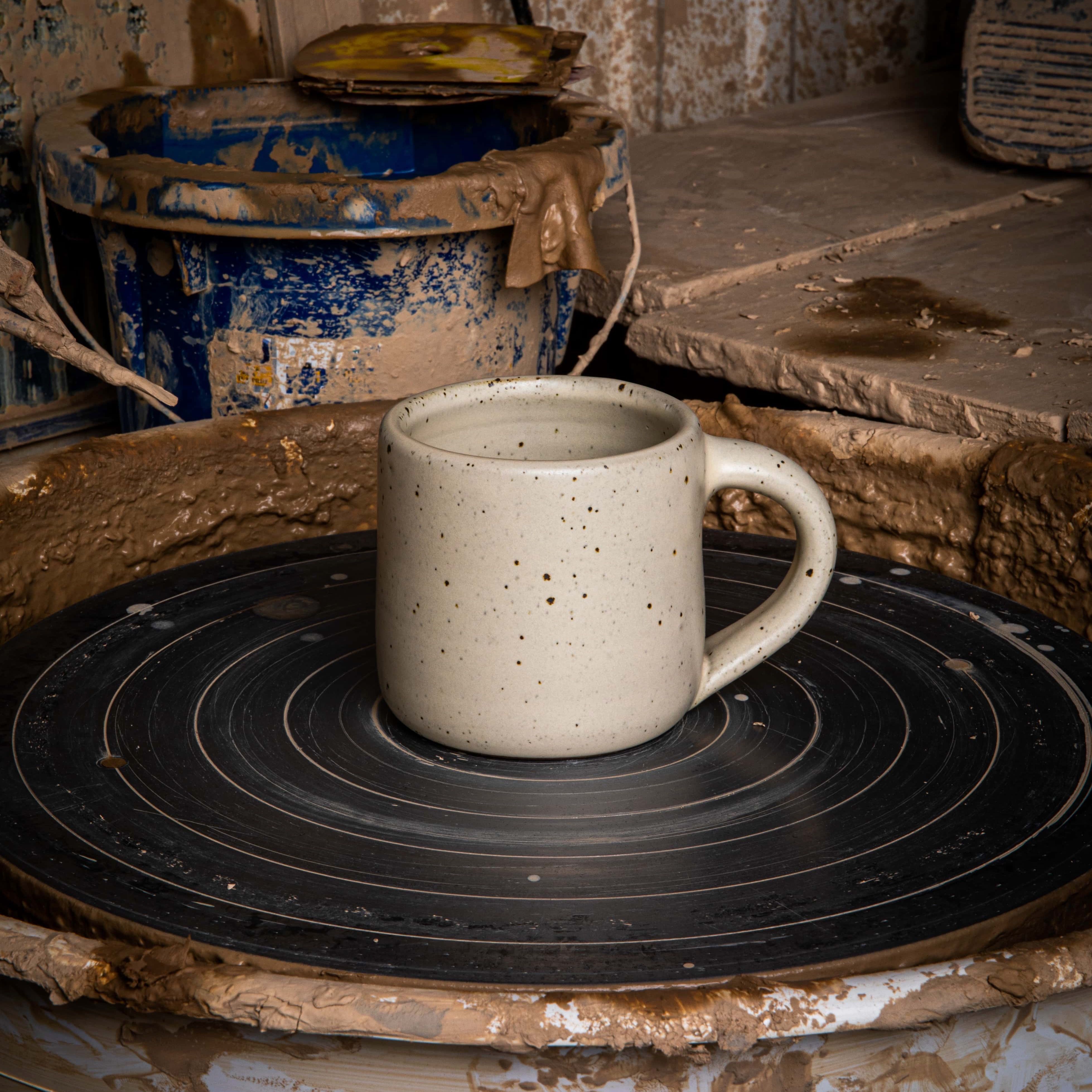 In a clay workshop environment, a medium sized ceramic mug with handle sits on a potter's wheel. The mug is in a warm, tan-toned, off-white color featuring iron speckles