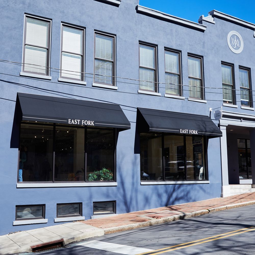 Outside view of East Fork Asheville, a light blue commercial building with black awnings over the windows and a second floor of windows with full blinds.