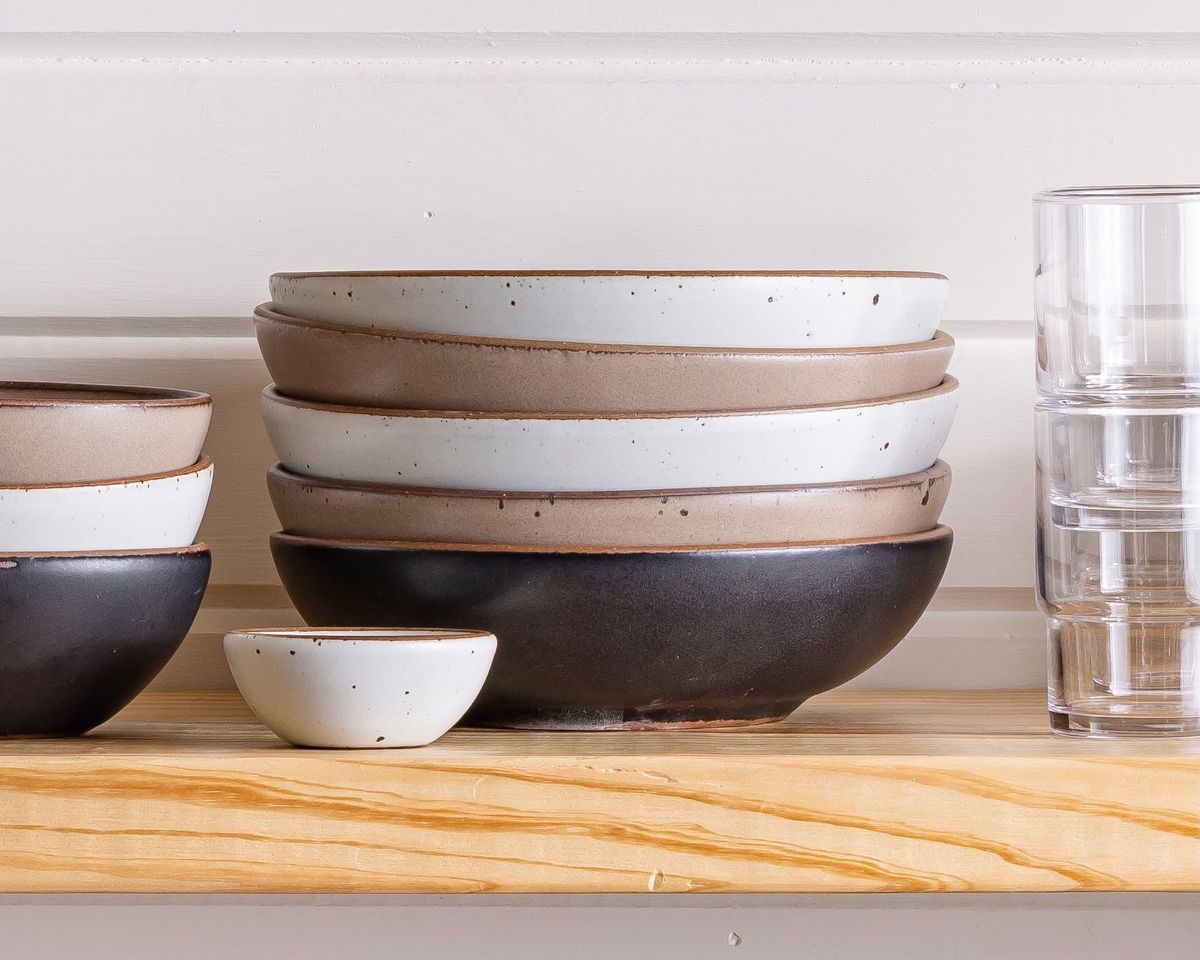 On a shelf is a stack of shallow ceramic bowls in cool white, neutral, and black colors. The bowls are surrounded by smaller sized bowls and a stack of glasses.