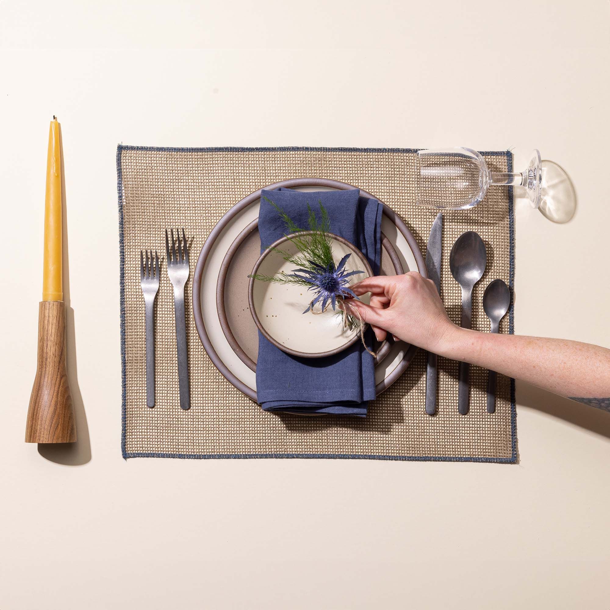 A hand arranging a place setting with 3 ceramic plates of different sizes in white, tan, and cream, steel flatware, a taper candle, and a wine glass.