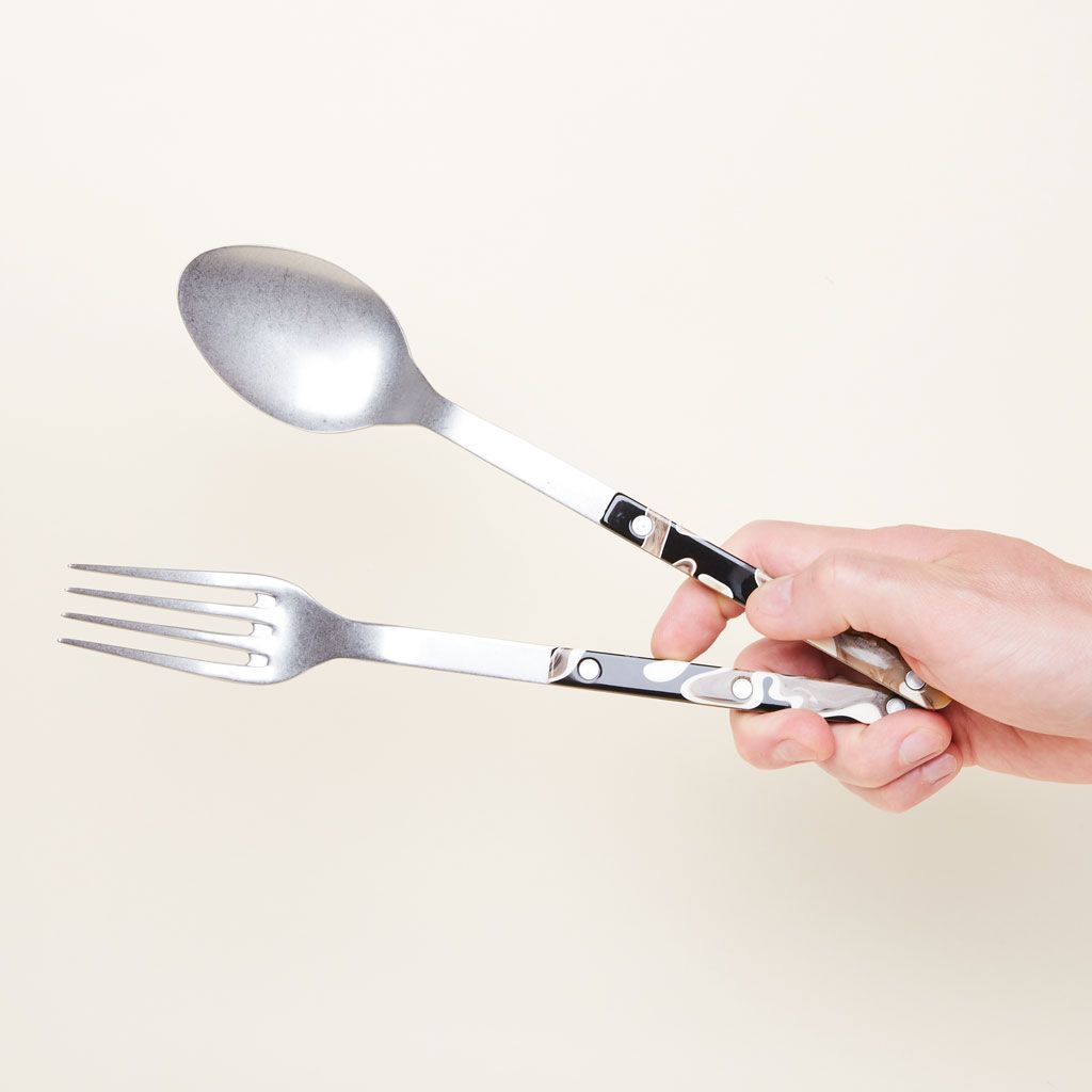 A serving fork and spoon in silver, black and brown held at a near-vertical angle by a hand