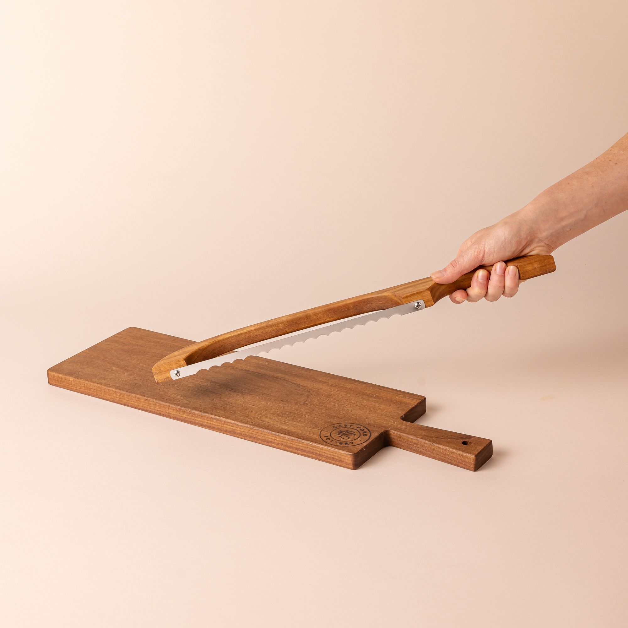 A wood rectangle bread board with an "East Fork Pottery" stamp on the corner and a handle. A hand holds a matching wood bread bow above it.