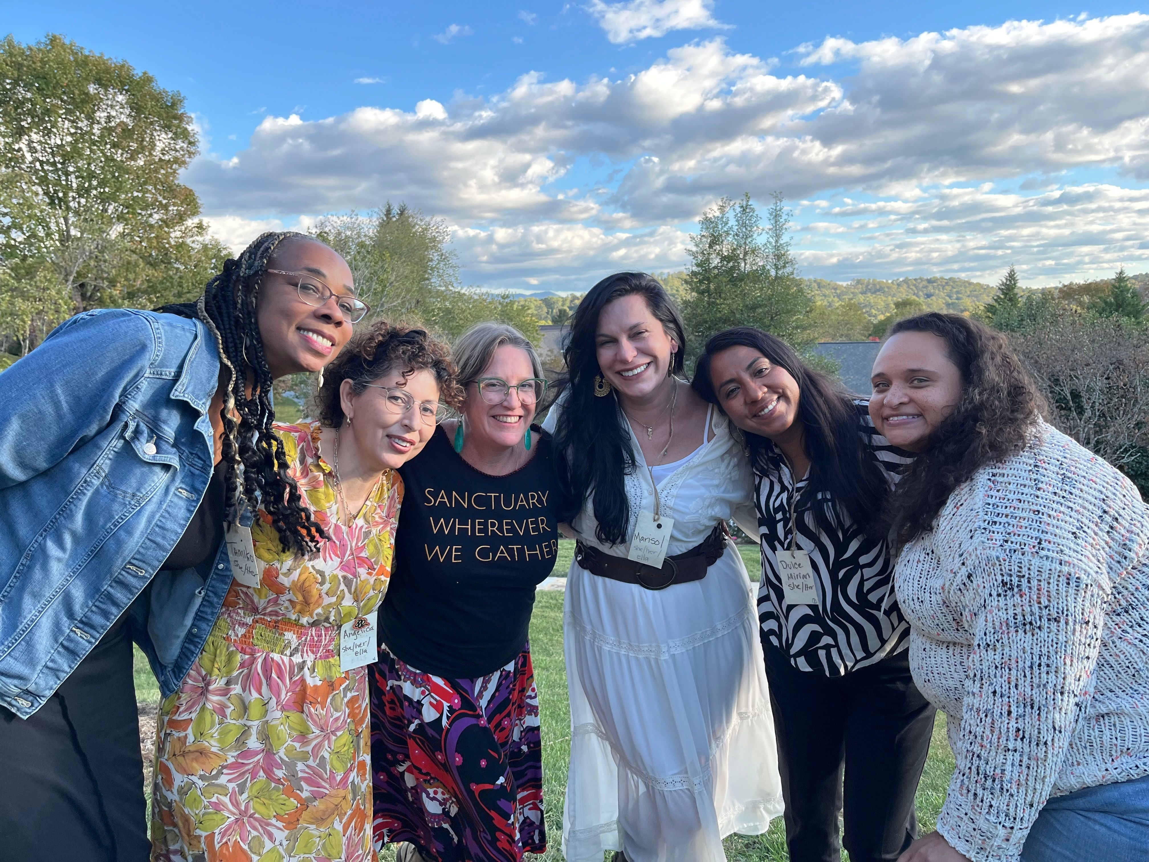 Six women smile with their arms around each other outdoors.