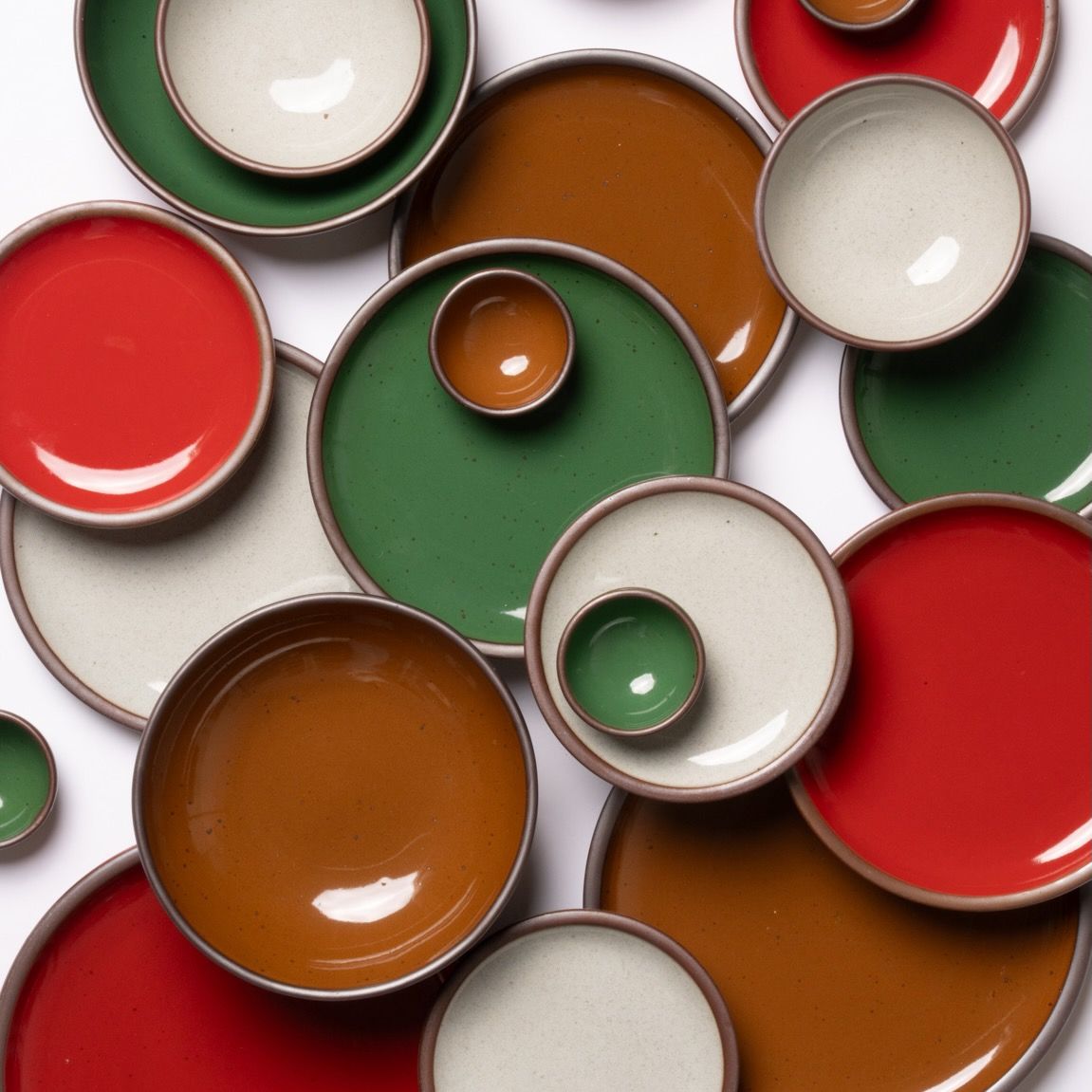 Overhead view of colorful ceramic plates and bowls in glossy red, green, brown, and icy white arranged in a layered, overlapping flatlay.