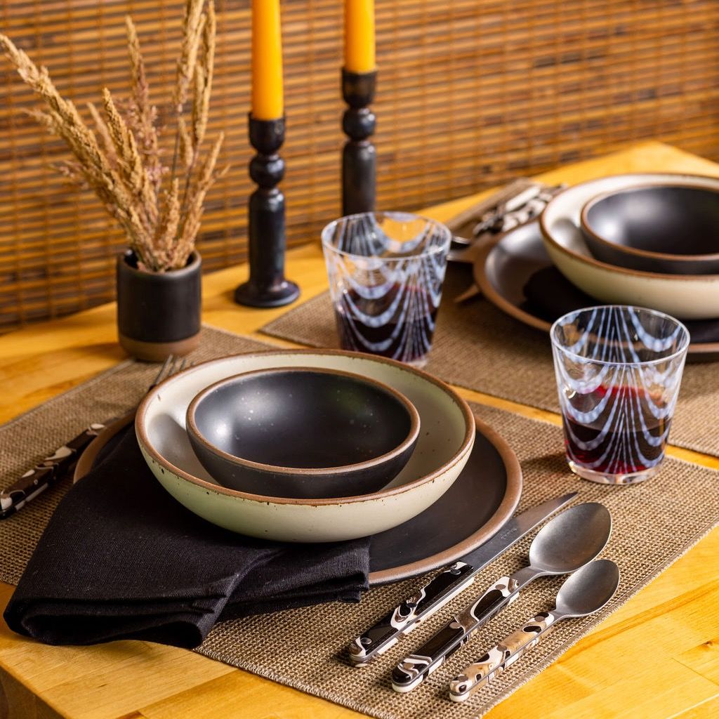 A wood dinner table setting with ceramic bowls and plates in black and cream, sophisticated marble flatware, rippled tumblers, dried grass arrangement, and tapered candles.
