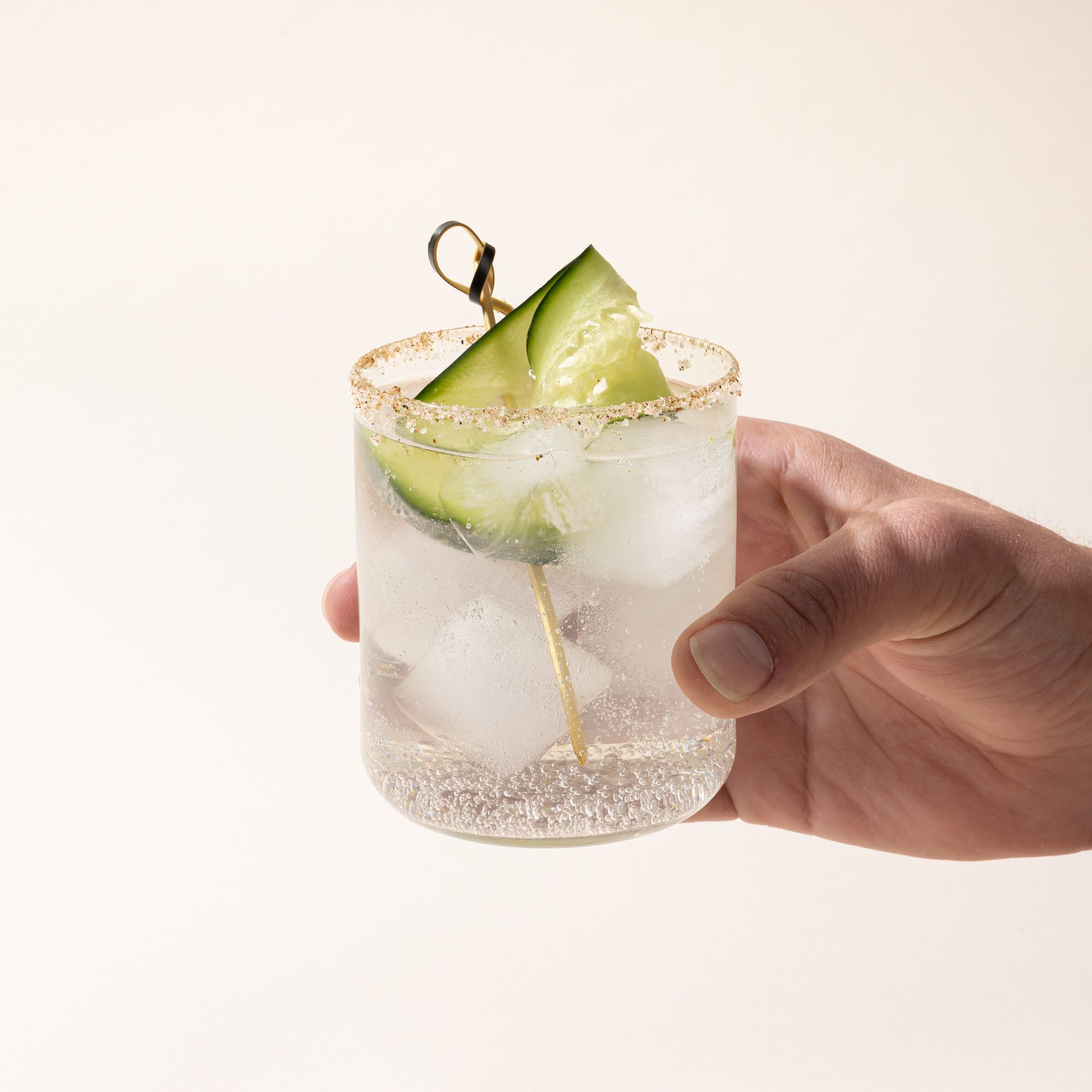 A hand holds a straight walled clear glass with a clear cocktail and cucumber garnish.