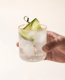 A hand holds a straight walled clear glass with a clear cocktail and cucumber garnish.