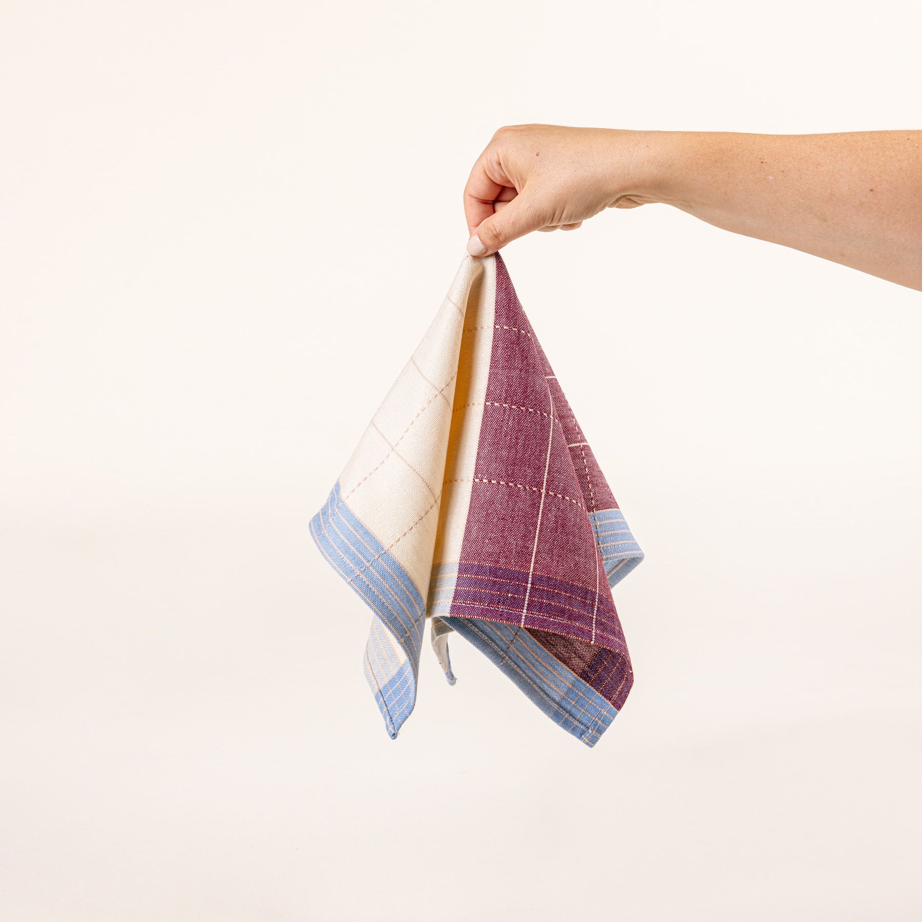 A hand holds the center of a flat plaid napkin in plum and periwinkle colors with white gridlines.