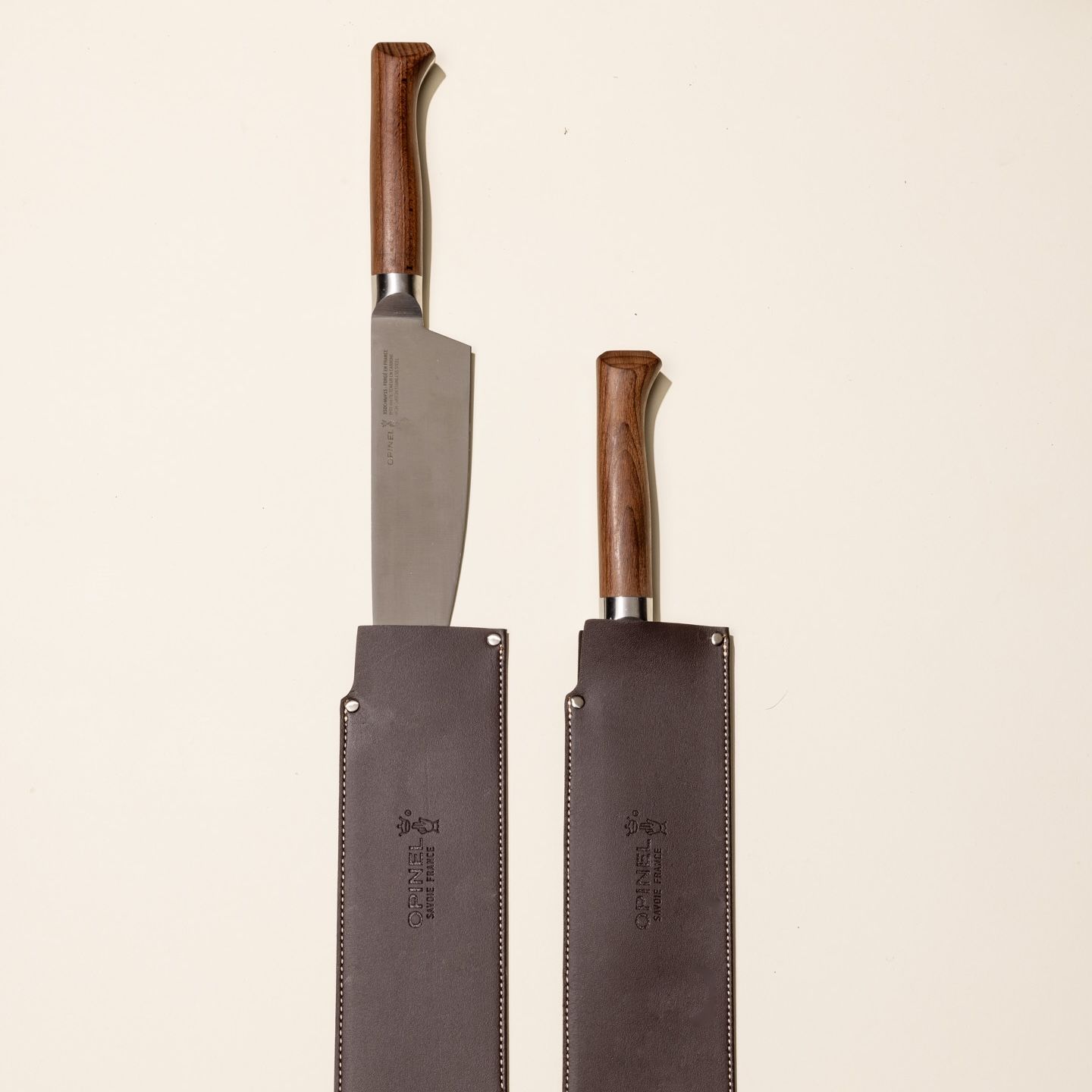 A chef's knife with a sharp steel blade and a beveled dark wood handle sits with a leather sleeve on the blade. Next to it is another knife sitting fully inside a leather sleeve.