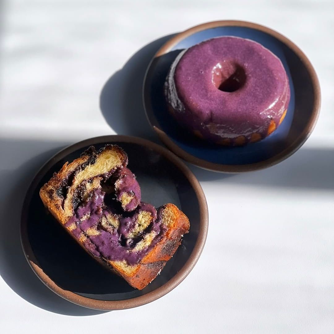 Slice of purple ube babka and a glazed ube bundt cake on black and medium blue ceramic dessert plates in natural light.