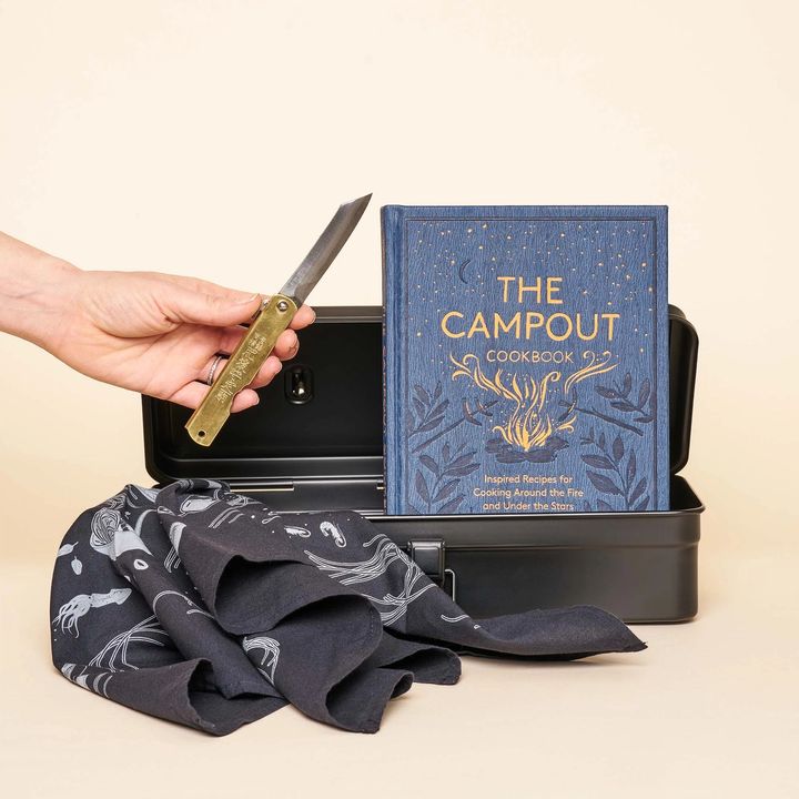 A steel box opens to display a navy blue cookbook ("The Campout Cookbook"). A hand holds a brass handled pocket knife with Japanese inscriptions on the handle. A black and white bandana sits in front of the box.
