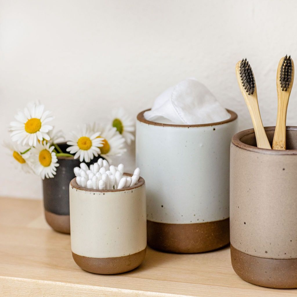 On a birch shelf, there are four candle vessels in various neutral colors, each holding something different: q-tips, toothbrushes, flowers. 