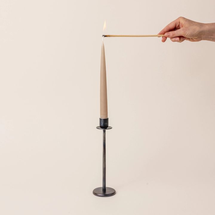 A hand is about to light a taupe taper candle sitting in a tall iron candlestick holder. The candlestick holder with a flat circle base and topped with a simple taper candle holder.