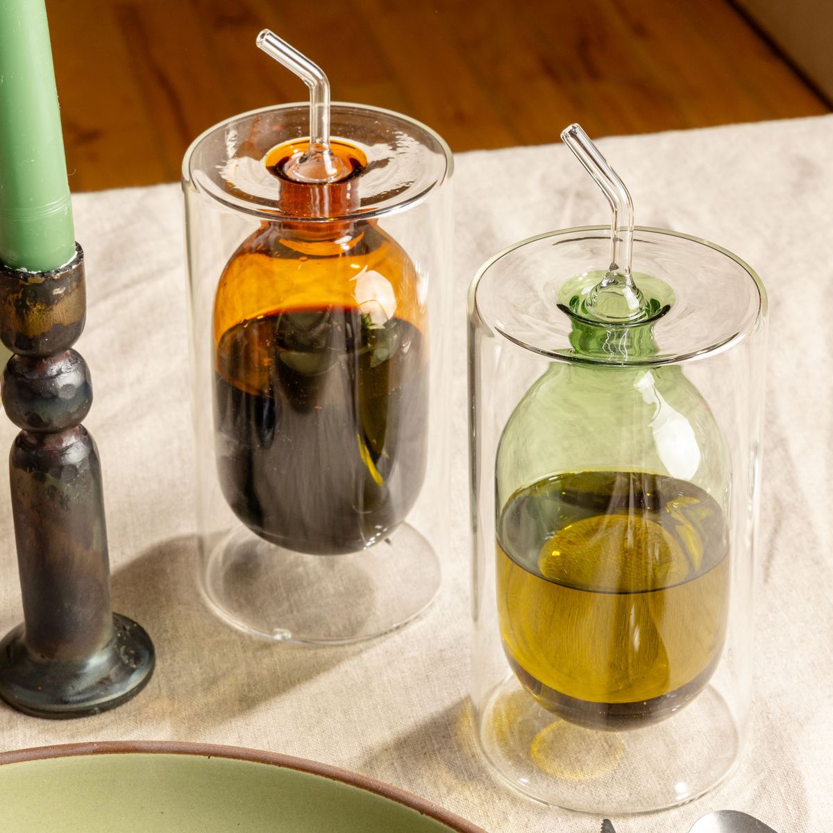 Two artful glass cruets in a an orange and light green color. There is a rounded bulb that acts as a container for the cruet, surrounded by a doubled glass wall.