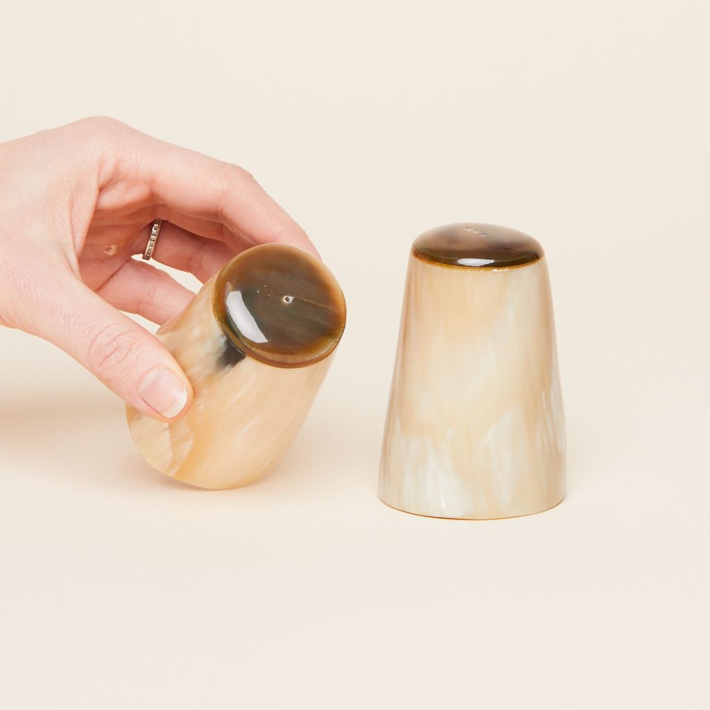 A hand tips a salt shaker toward the camera while its matching pepper shaker stands to its right