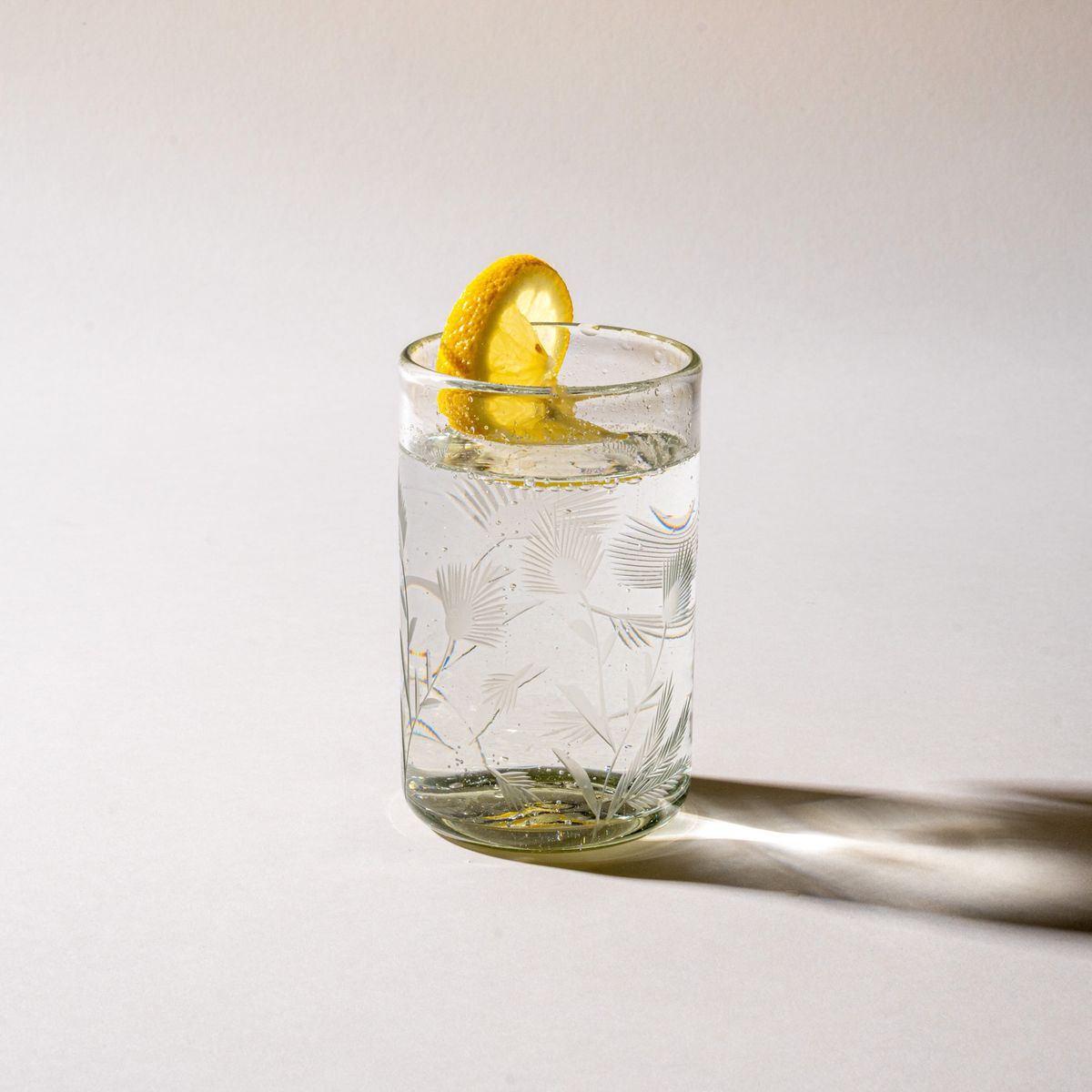 A clear tumbler glass made of recycled glass with etched thistle designs on it filled with water and topped with a slice of lemon.