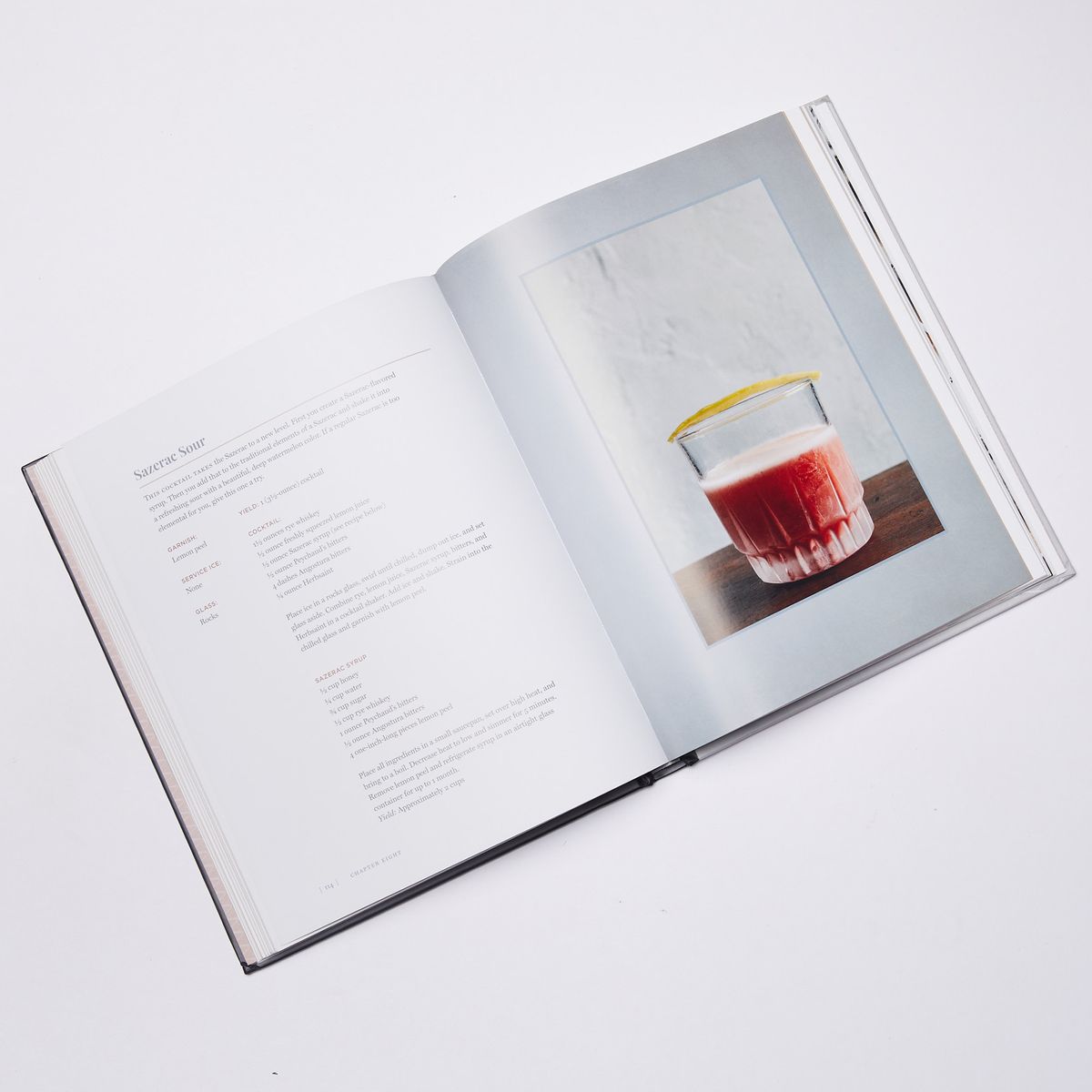 Open cookbook with a recipe for a sazerac sour on the left with a photo of a bright red cocktail in a frosty tumblr on the right