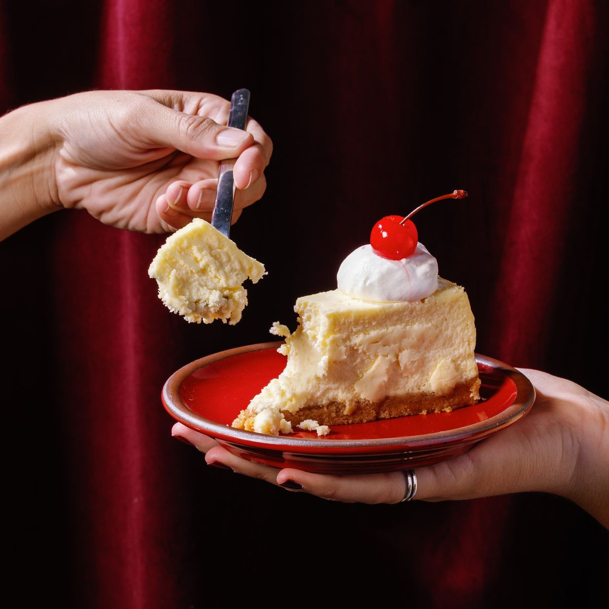 A hand holds out a small dessert plate in a bold glossy red color, with a piece of cheesecake and a hand holding a fork grabbing a bite.