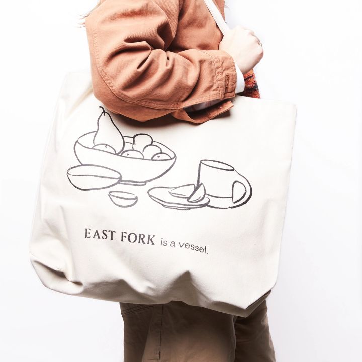 Person holding a cream canvas tote on their shoulder with their hand holding onto the handle - the tote has a charcoal illustration on the front of a bowl with fruit, little bowls, plate, and a mug. Underneath, it reads 'East Fork is a vessel'.