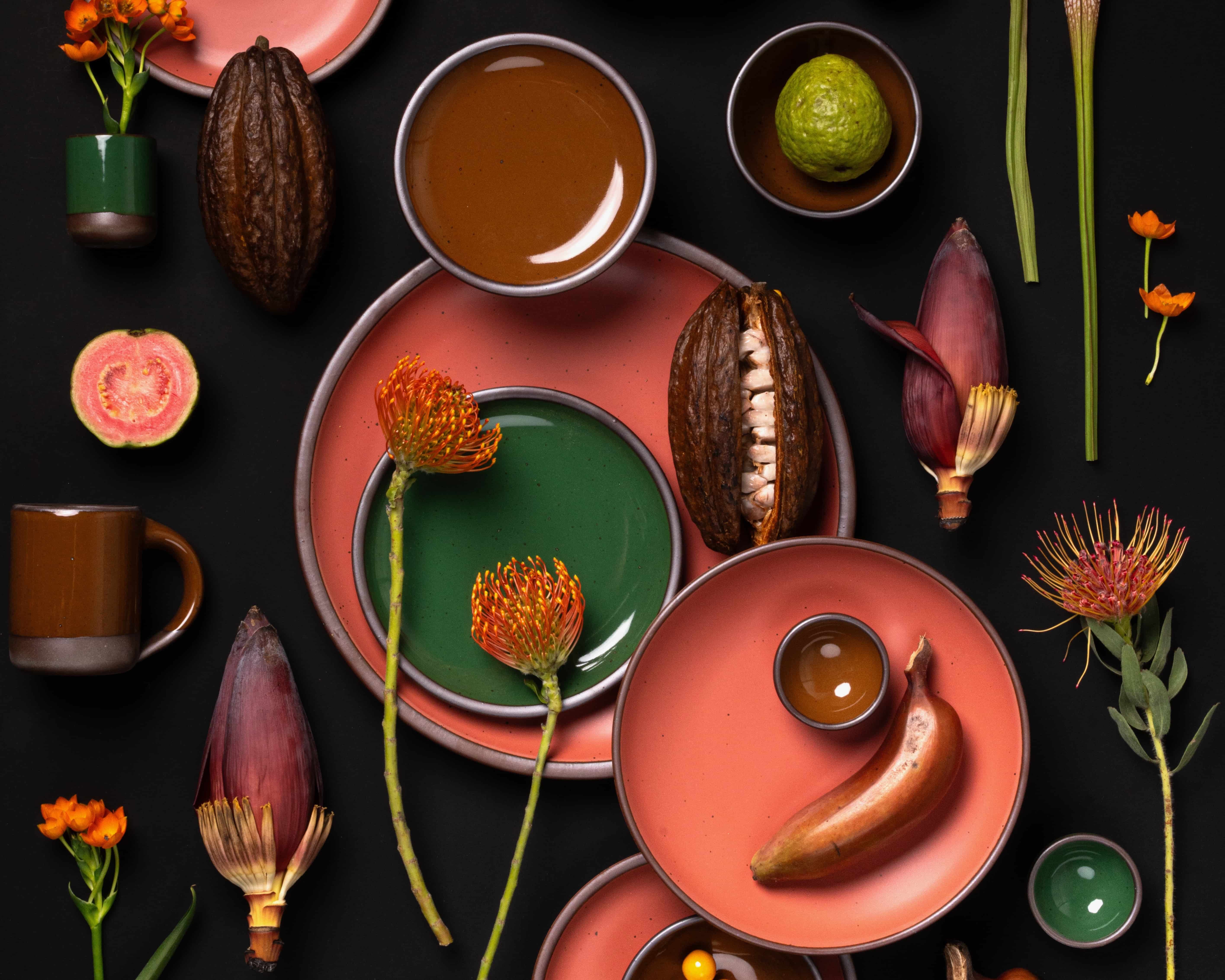 Colorful flat lay with coral, glossy green, and glossy brown plates styled with cacao pods, guava, banana blossoms, and orange flowers.