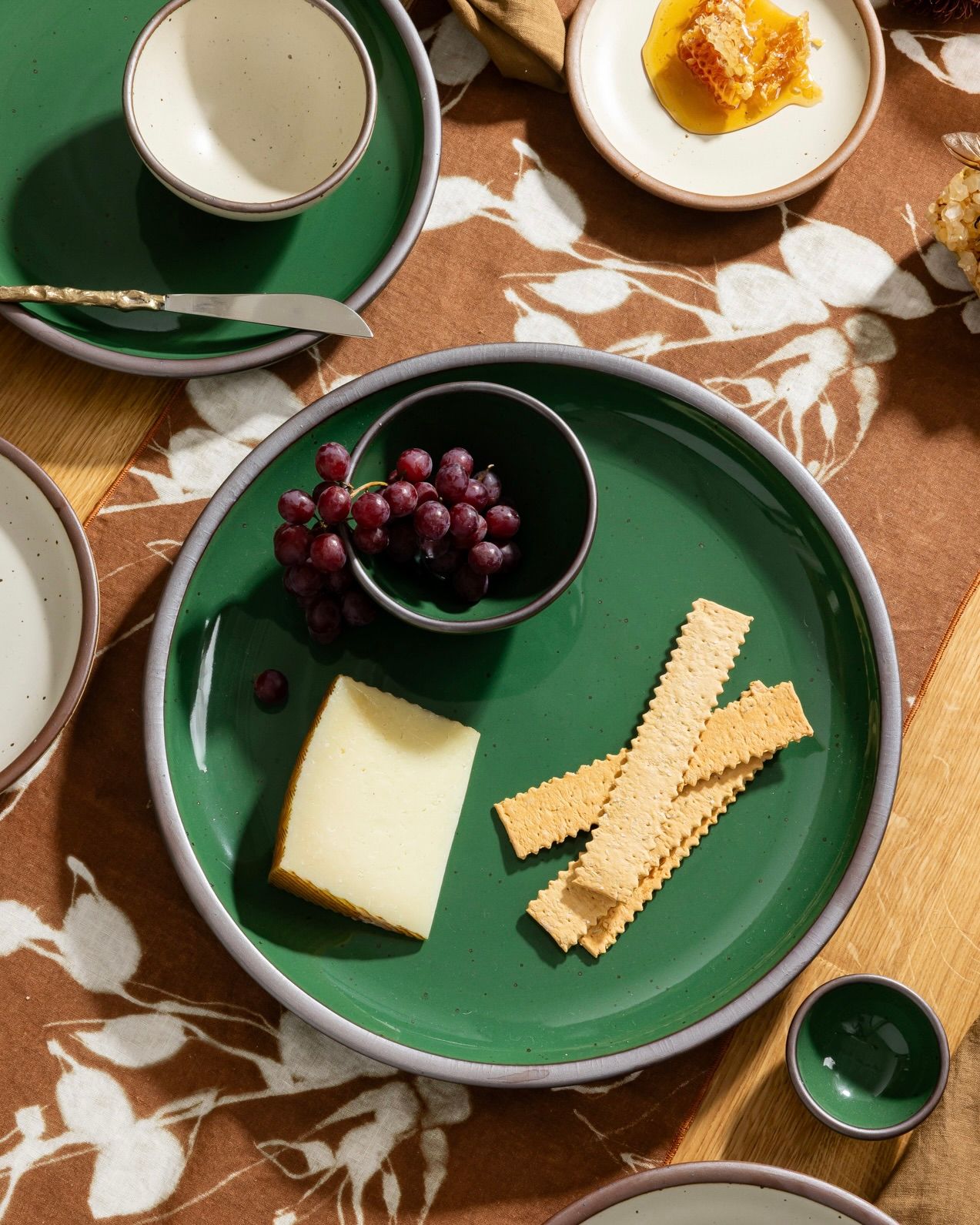 Glossy emerald green plate with grapes, cheese, and crackers beside honeycomb on a patterned brown tablecloth.