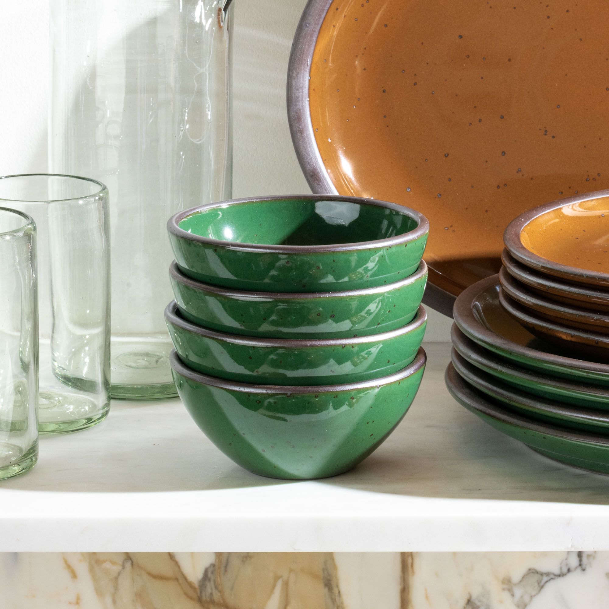 A stack of 4 small dessert sized ceramic bowls in a glossy emerald green color on a white shelf