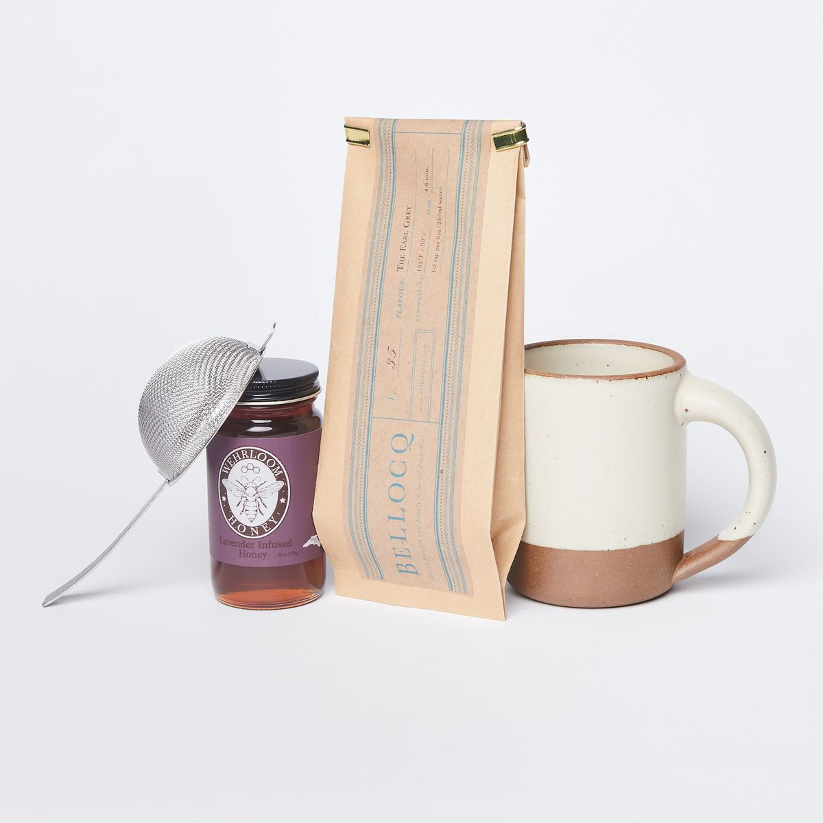 Tea strainer leaning on a jar of honey, brown bag of tea with turquoise lettering "Bellocq", panna cotta mug