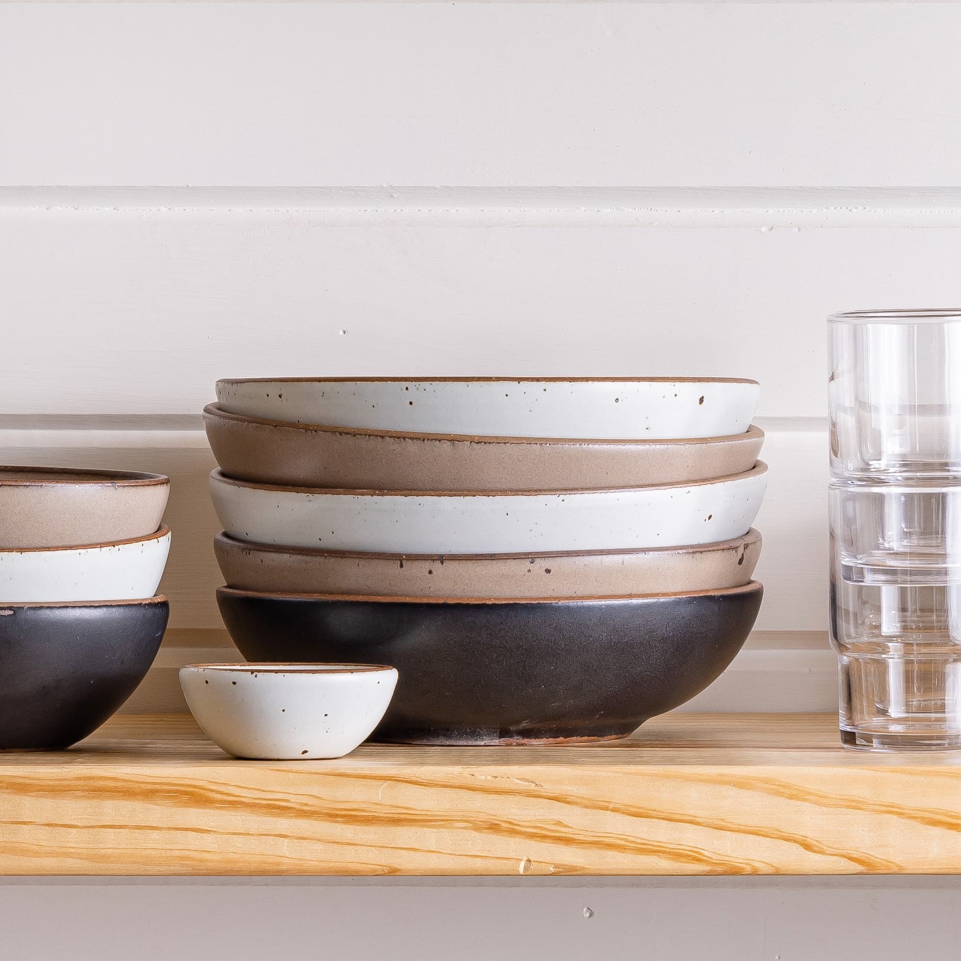 On a shelf is a stack of shallow ceramic bowls in cool white, neutral, and black colors. The bowls are surrounded by smaller sized bowls and a stack of glasses.