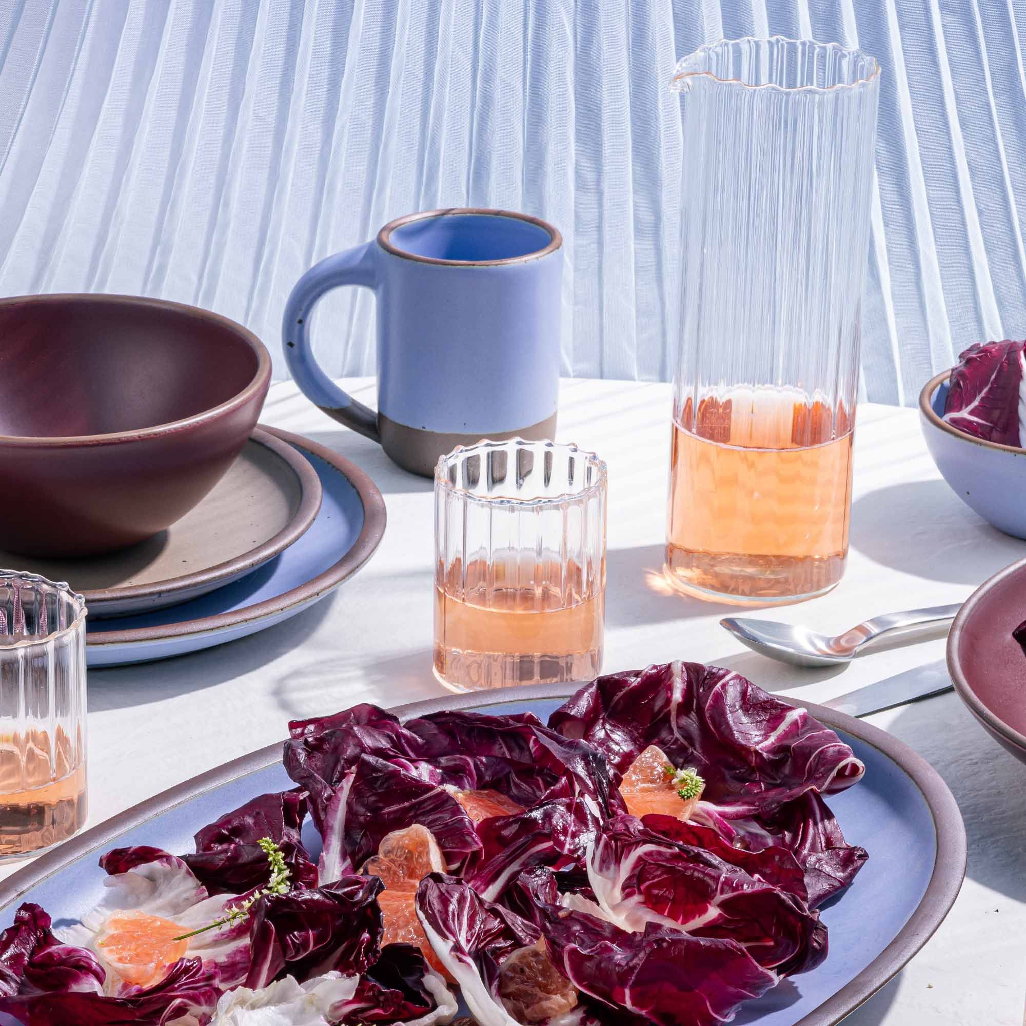 Fluted pitcher and short tumblers sit with periwinkle ceramic pots and plates with salad