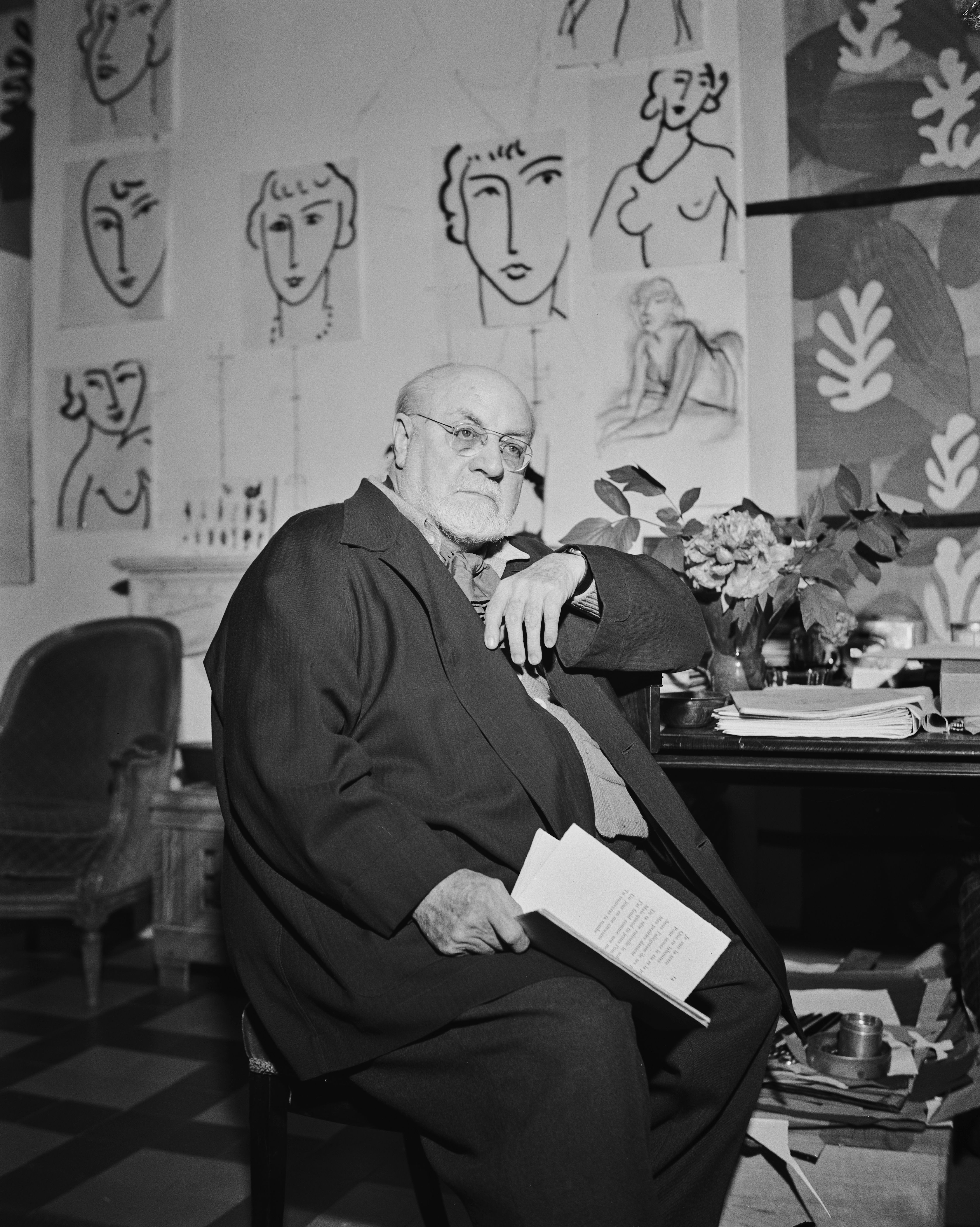 Black and white photograph of Henri Matisse sitting in his studio holding a paper and a large wall of his artwork behind him.