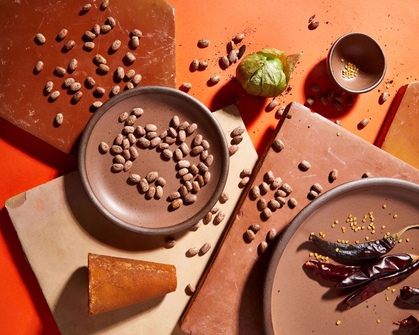 An overhead view of plates and tiles in a warm brown color with ingredients like dried pinto beans, peppers, seeds, and a tomatillo