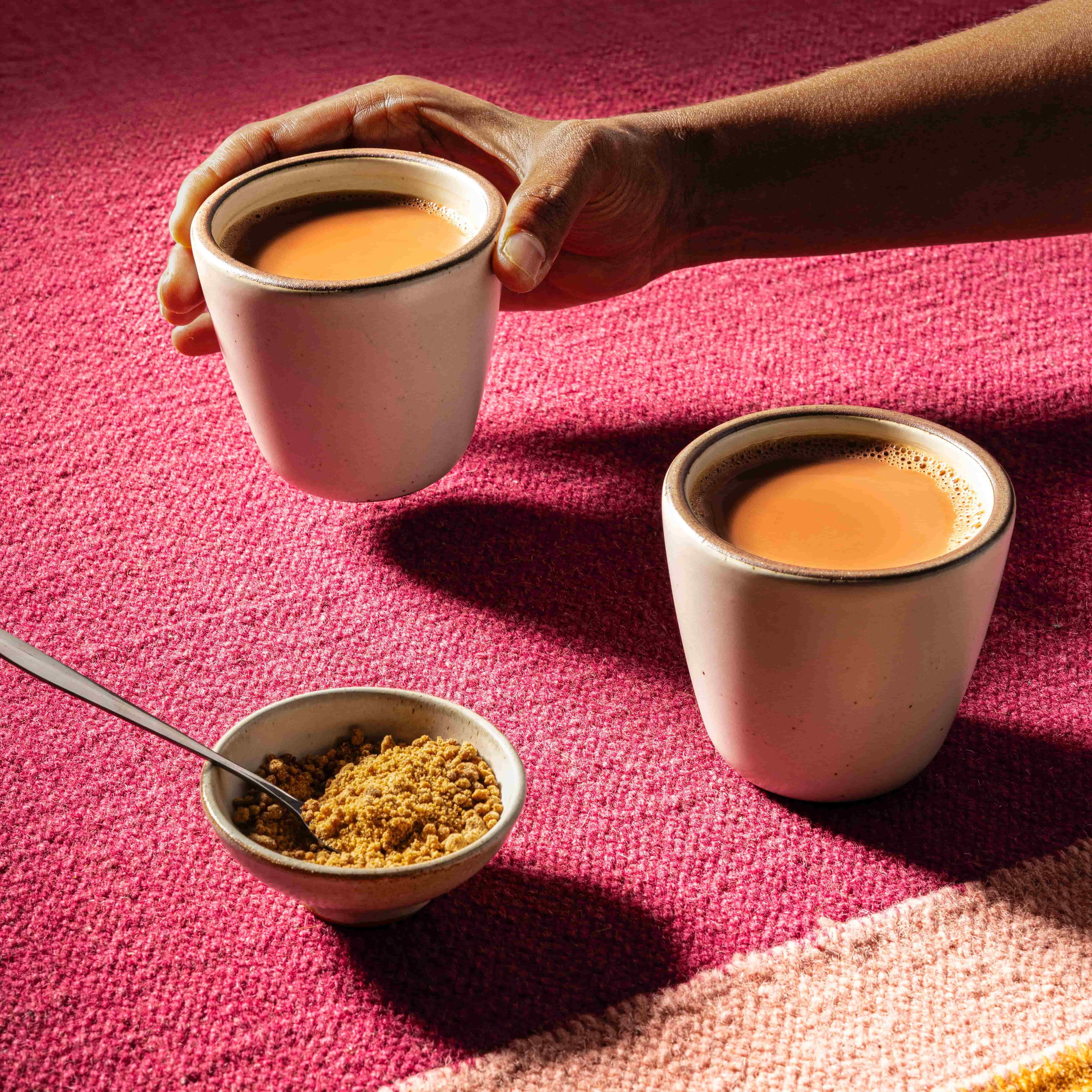 A hand holds out a ceramic cup with another one next to it, both filled with chai, along with a small bowl of jaggery, against a pink textile