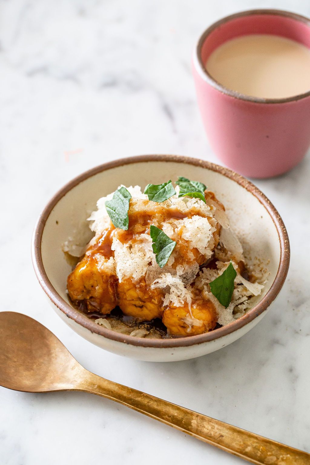 Caramelized plantains in an off-white ceramic bowl with a pink cup of chai and brass spoon