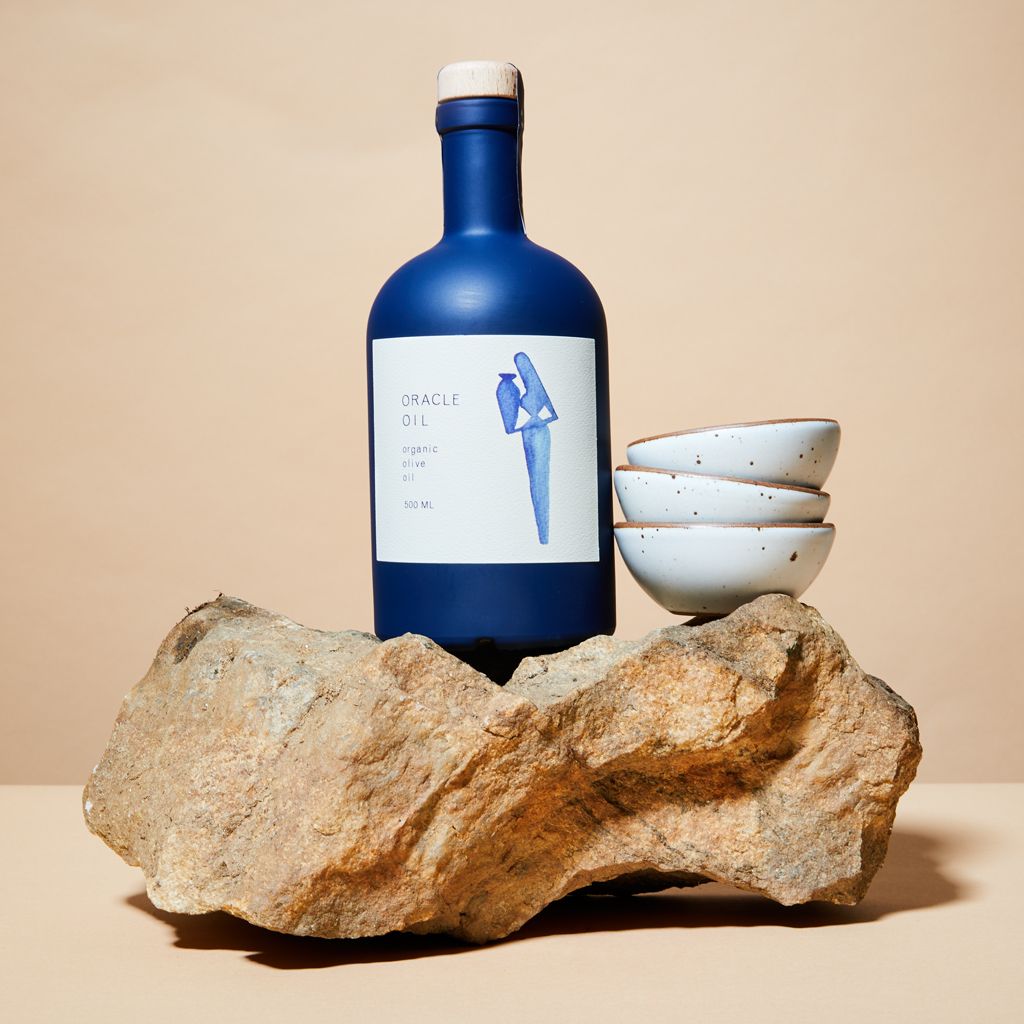 A Grecian blue bottle of olive oil with with a white label and cork cap, sitting on top of a granite boulder next two three eggshell bitty bowls stacked on top of one another.